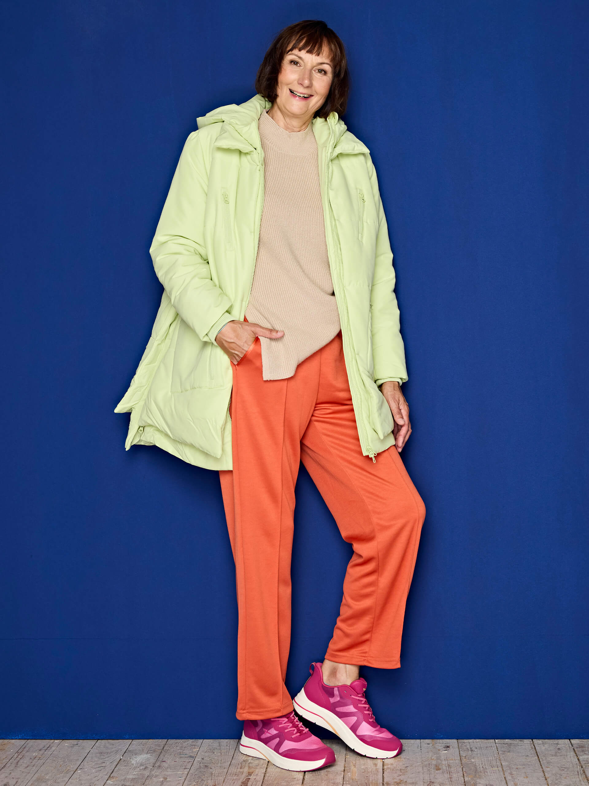 Eine Frau trägt eine helle, wattierte Jacke, einen beigen Pullover, orangefarbene Hosen und pinke Sportschuhe vor blauem Hintergrund.