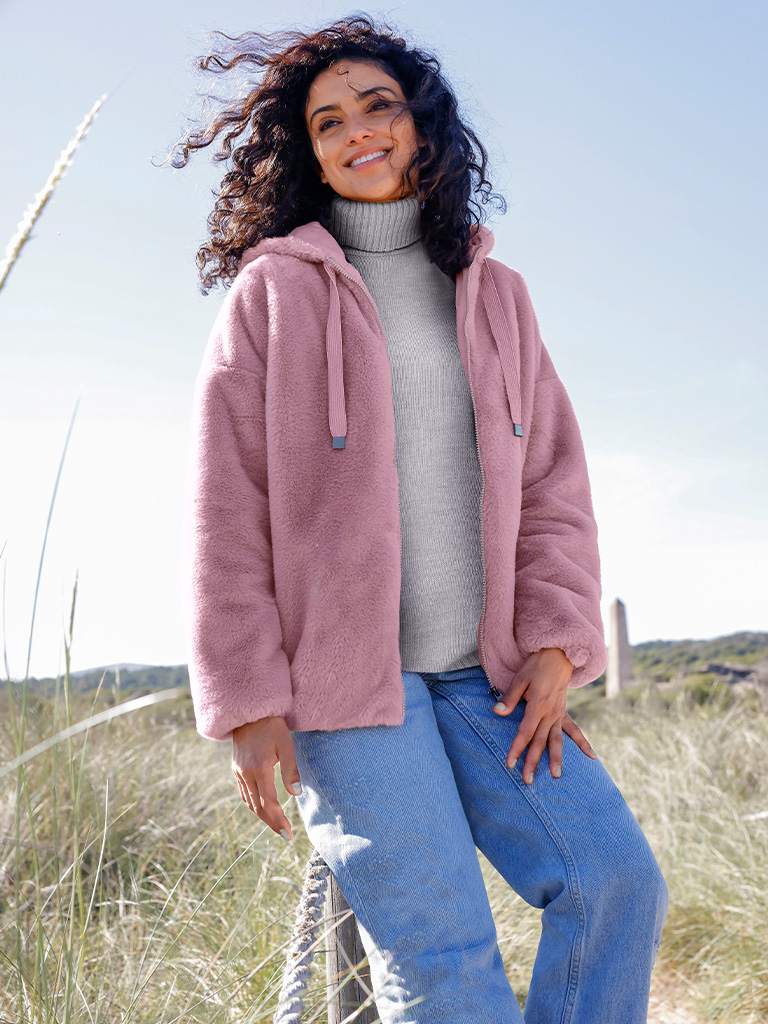 Ein Model trägt eine mauvefarbene Teddyfelljacke, einen grauen Rollkragenpullover und blaue Jeans, steht draußen in der Natur.