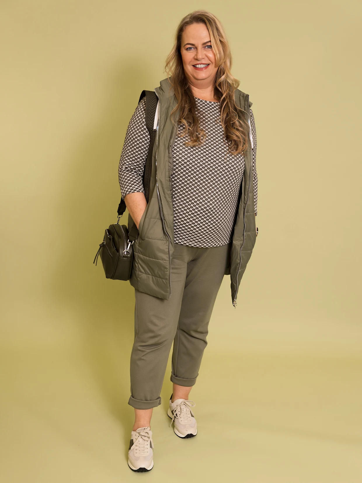 Eine blonde Frau trägt eine khakifarbene Longweste, ein oliv-gemustertes Shirt, khakifarbene Jogg Pants, eine dunkle Handtasche und beige-khakifarbene Sneaker. Der Hintergrund ist blassgelb.