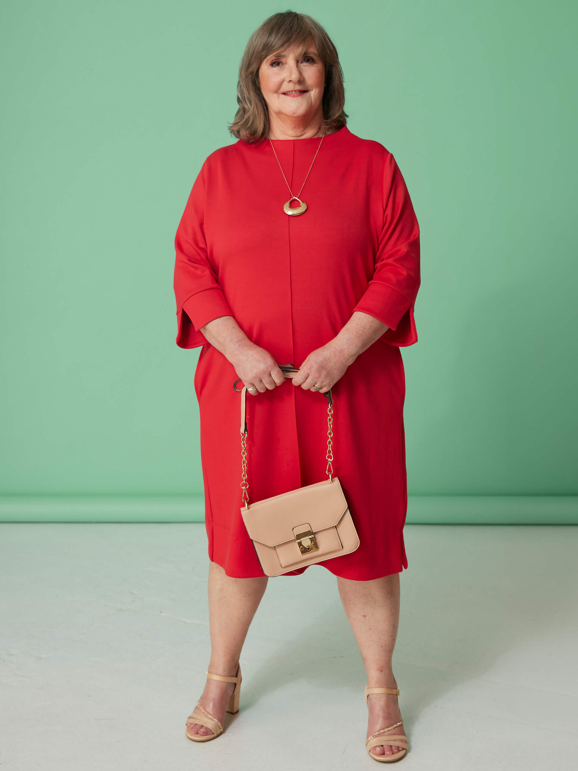 Ein Model trägt ein rotes Kleid und Sandalen, hält eine beigefarbene Umhängetasche und steht vor mintgrünem Hintergrund.