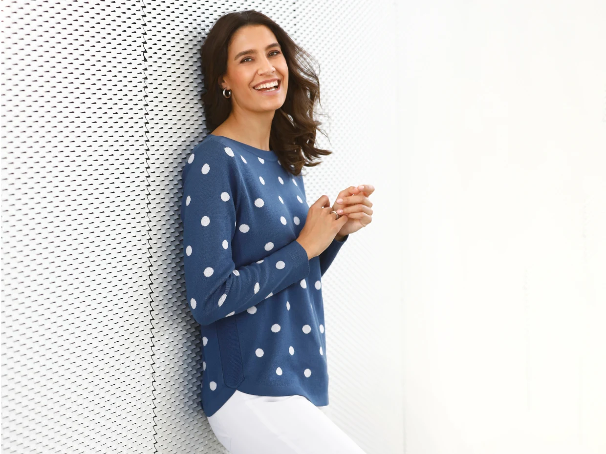 Frau trägt ein blaues Langarmshirt mit weißen Punkten und eine weiße Hose, lehnt an einer perforierten weißen Wand.