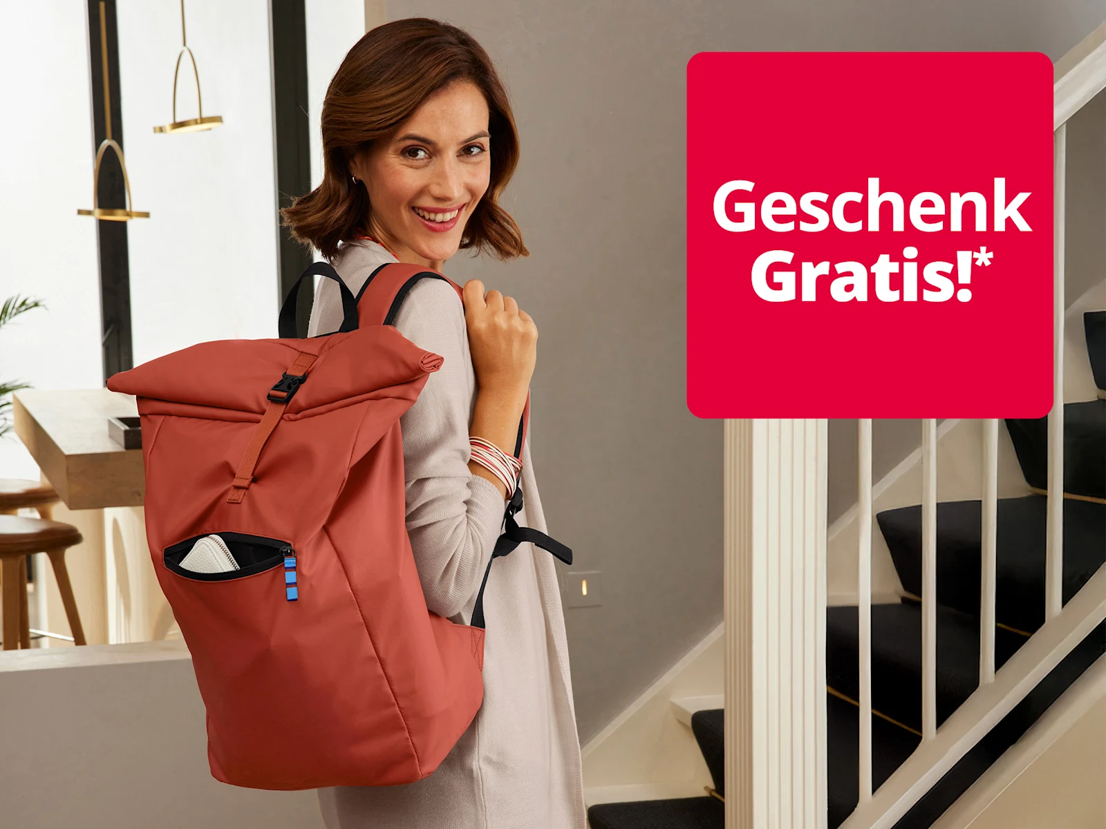 Das Bild zeigt eine Frau mit hellbraunen Haaren, die einen roten Rucksack trägt, in einem modernen, hellen Flur mit Treppe und eleganter Einrichtung. Auf einem roten Quadrat steht der Text 'Gratis Versand & Geschenk!*' in weißer Schrift. Das Bild zeigt eine Frau mit hellbraunen Haaren, die einen roten Rucksack trägt, in einem modernen, hellen Flur mit Treppe und eleganter Einrichtung. Auf einem roten Quadrat steht der Text 'Gratis Versand & Geschenk!*' in weißer Schrift.