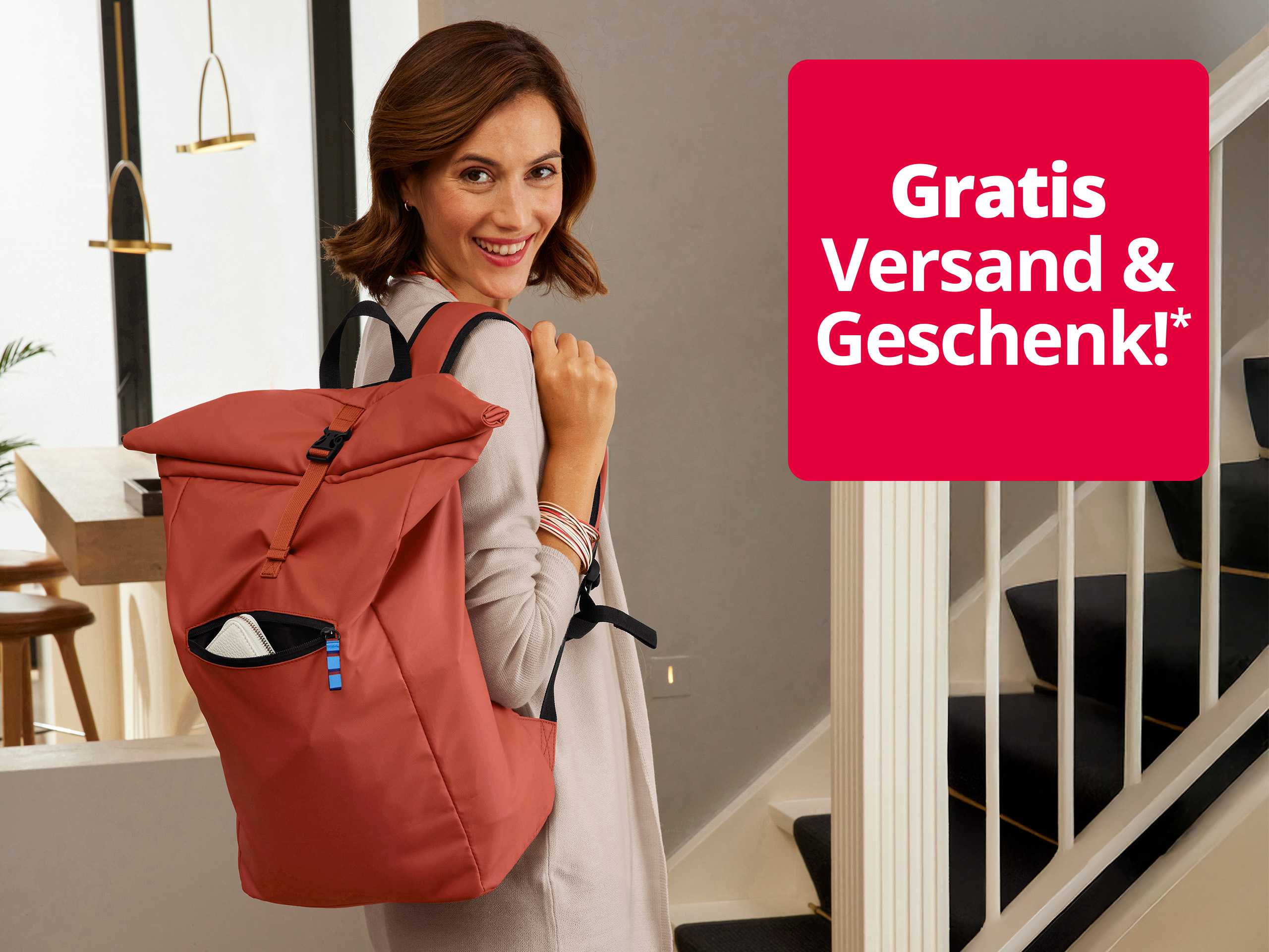Das Bild zeigt eine Frau mit hellbraunen Haaren, die einen roten Rucksack trägt, in einem modernen, hellen Flur mit Treppe und eleganter Einrichtung. Auf einem roten Quadrat steht der Text 'Gratis Versand & Geschenk!*' in weißer Schrift.