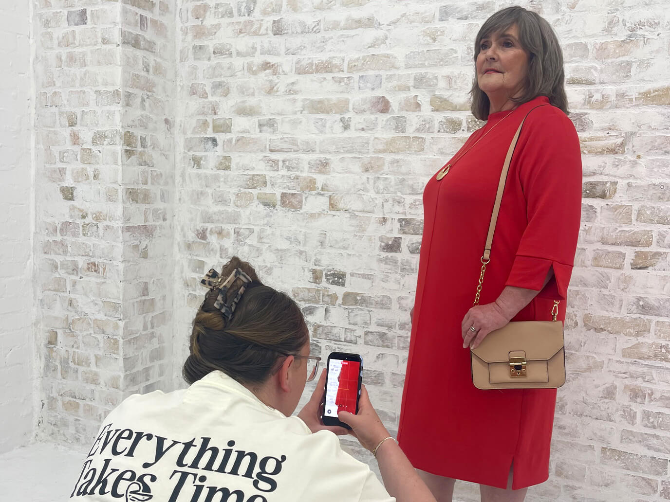 Ein Frau im roten Kleid steht vor einer weißen Backsteinwand, während eine andere Frau sie mit einem Smartphone fotografiert.