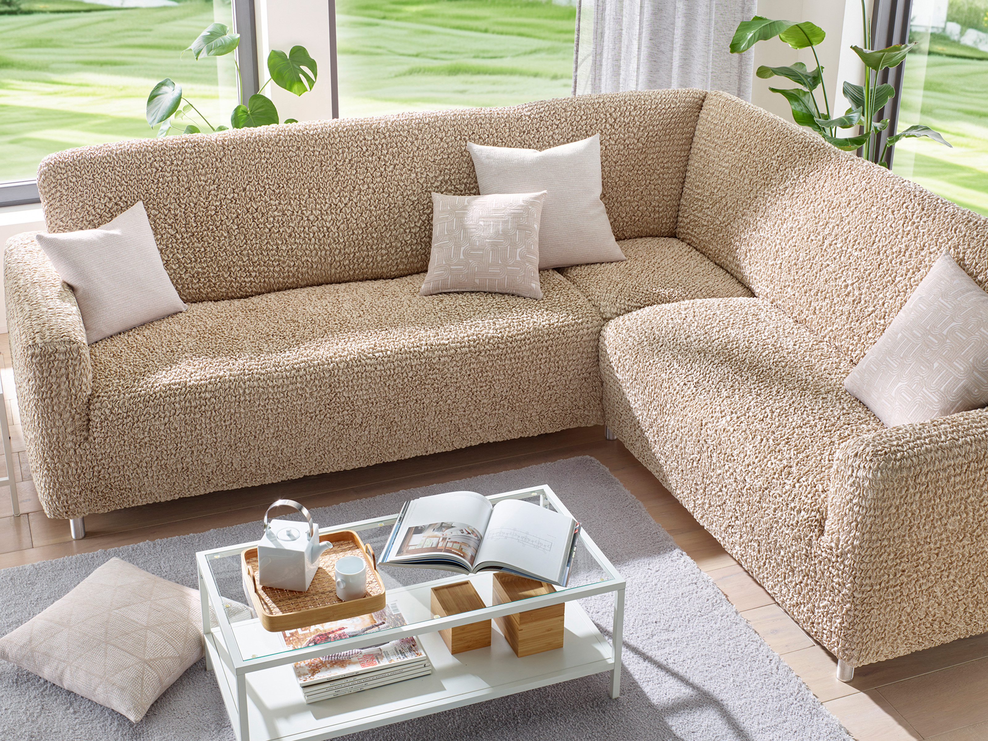 Ecksofa mit beigem, strukturiertem Bezug und hellen Kissen, davor ein Glastisch mit Büchern und Teekanne, helle Wohnzimmereinrichtung.