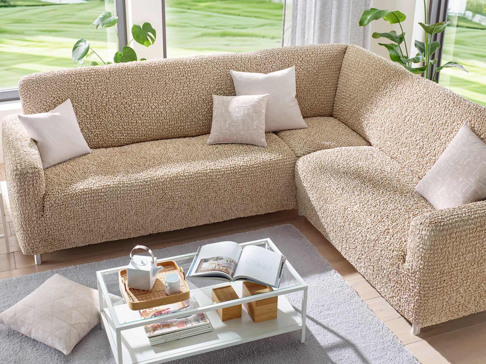Ecksofa mit beigem, strukturiertem Bezug und hellen Kissen, davor ein Glastisch mit Büchern und Teekanne, helle Wohnzimmereinrichtung.