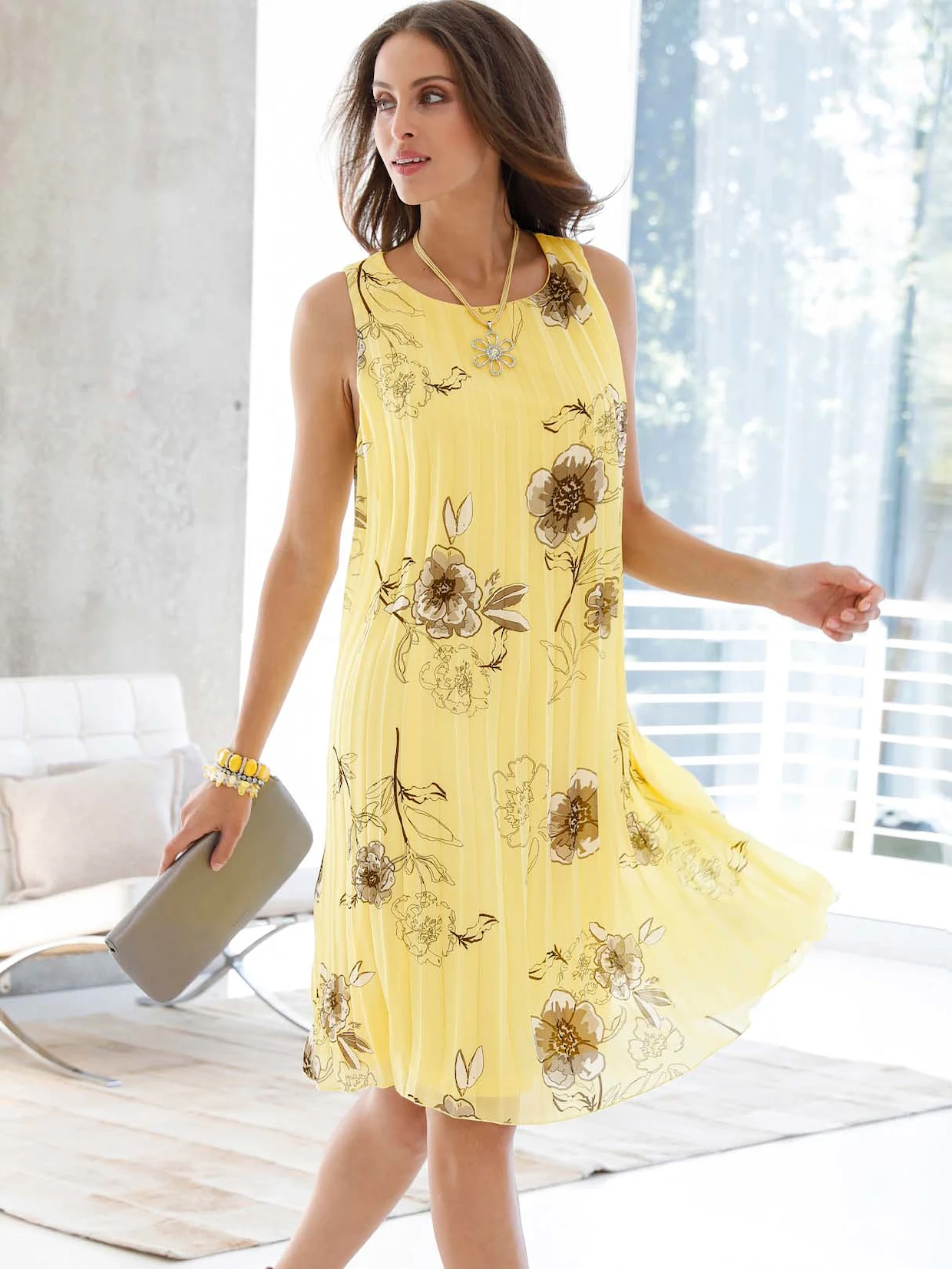 Frau trägt gelbes Sommerkleid mit floralen Mustern, kombiniert mit einer goldenen Halskette und einer grauen Clutch.