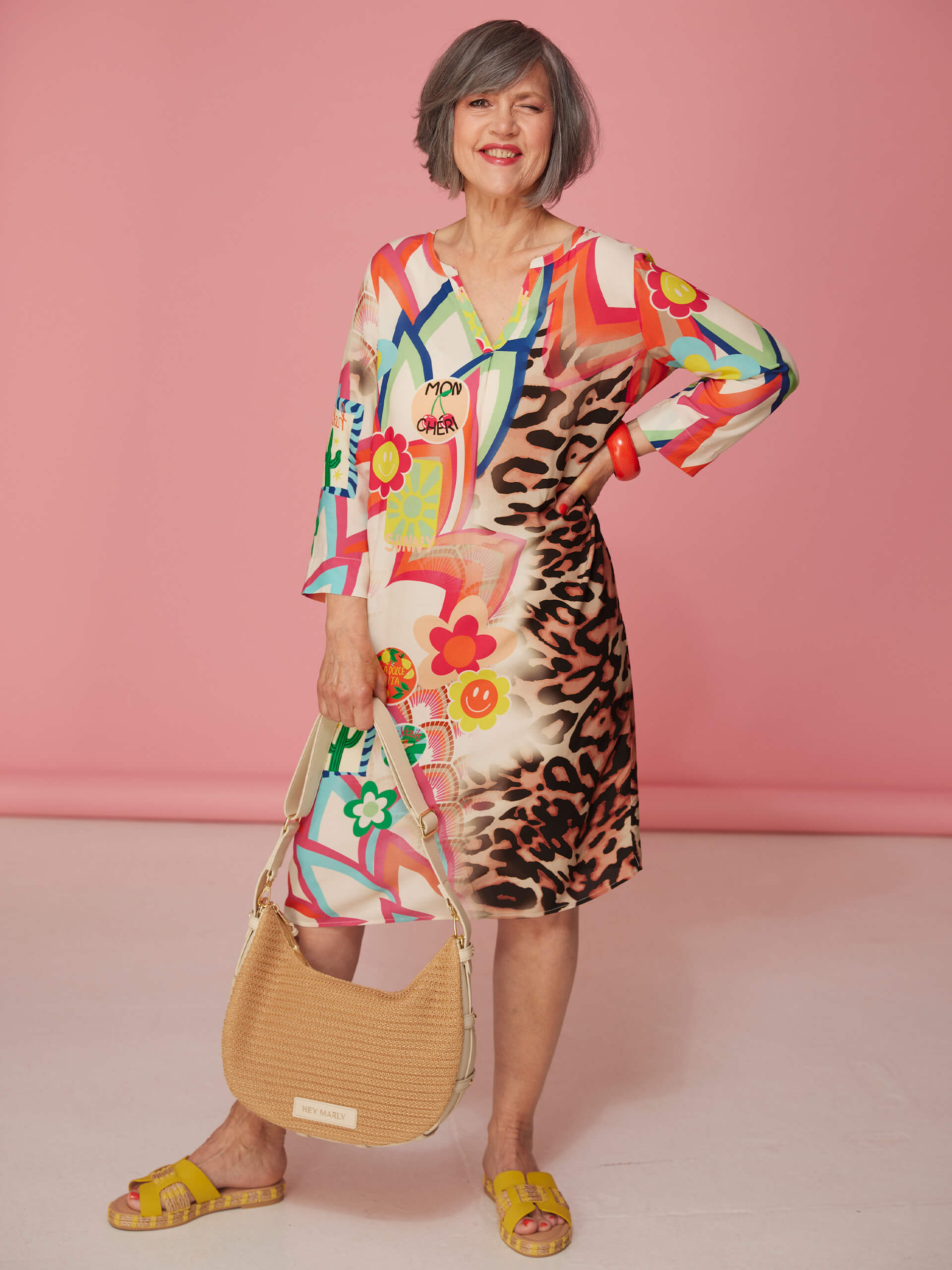 Ein Model trägt ein bunt gemustertes Kleid mit Leopardenmuster, gelbe Sandalen und hält eine geflochtene Tasche vor rosa Hintergrund.