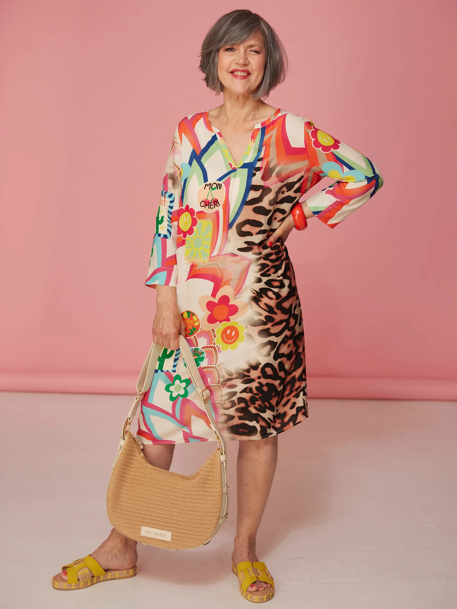Ein Model trägt ein bunt gemustertes Kleid mit Leopardenmuster, gelbe Sandalen und hält eine geflochtene Tasche vor rosa Hintergrund.