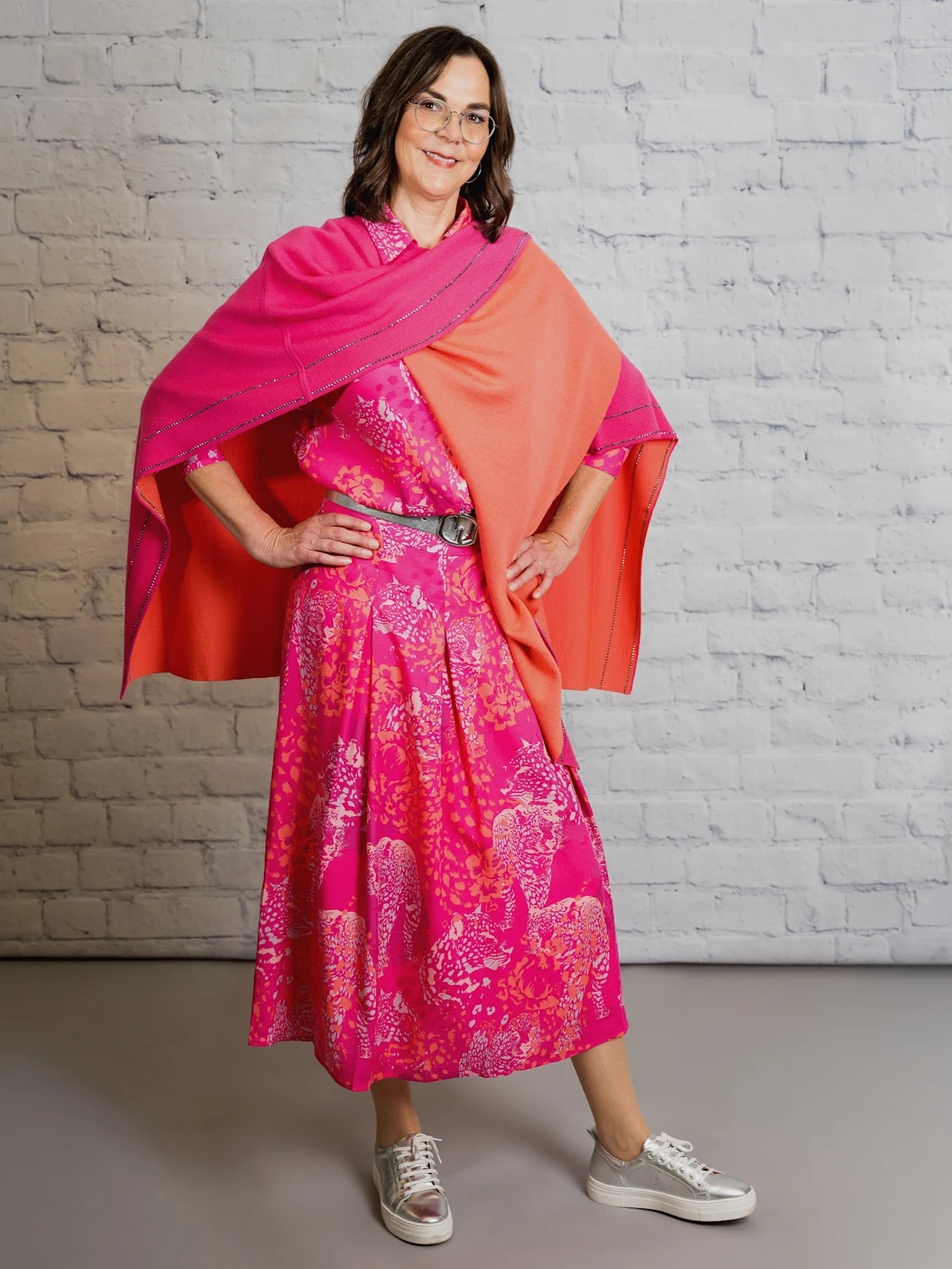 Eine dunkelhaarige Frau trägt eine fuchsia-koralle-bedruckte Bluse in Lyocell-Qualität, einen fuchsia-koralle-bedruckten A-Linien-Rock in TENCEL-Qualität, einen fuchsia-korallefarbenen Strickponcho mit Wende-Funktion, einen silberfarben Gürtel und silberfarbene Sneaker. Der Hintergrund ist eine weiße Wand.