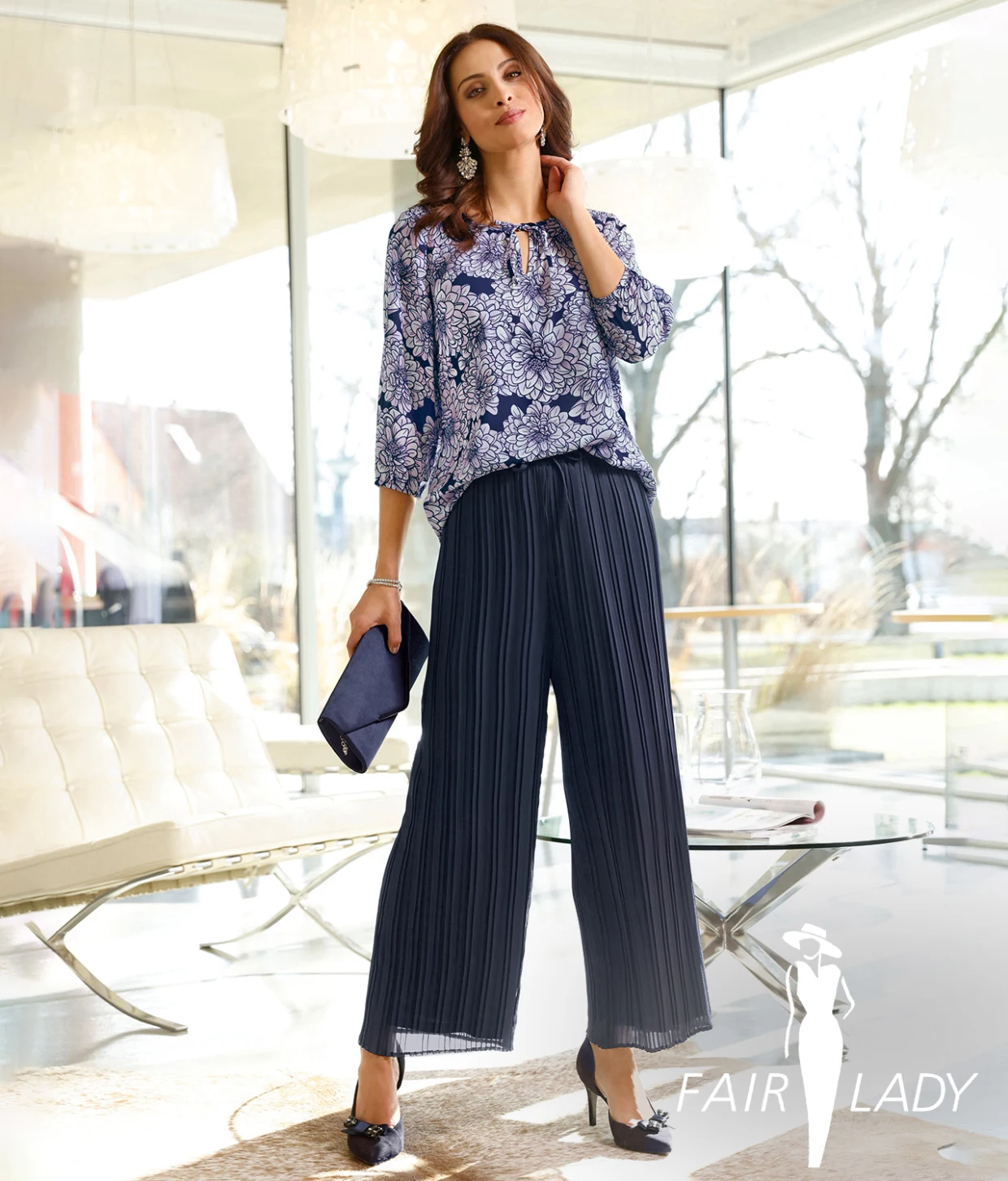 Frau trägt ein elegantes Outfit bestehend aus einer blau gemusterten Bluse und einer weit geschnittenen, plissierten Hose in Dunkelblau, kombiniert mit passenden High Heels und einer dunkelblauen Clutch. Im Hintergrund modernes Wohnzimmer mit großen Fenstern.