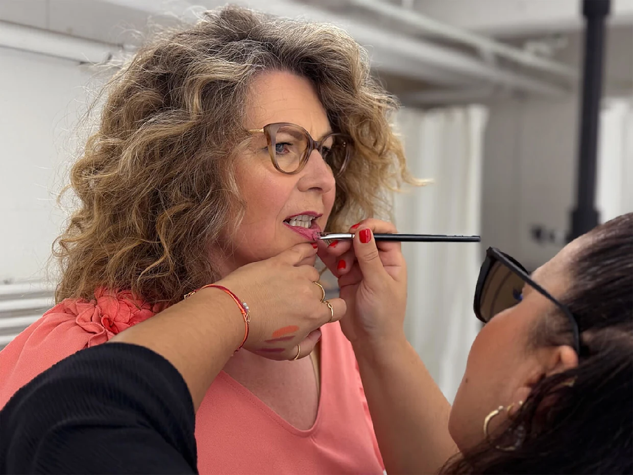 Eine Frau mit lockigem Haar wird geschminkt. Eine Person trägt Make Up auf das Gesicht der Frau auf. Der Hintergrund ist unscharf. Eine Frau mit lockigem Haar wird geschminkt. Eine Person trägt Make Up auf das Gesicht der Frau auf. Der Hintergrund ist unscharf.