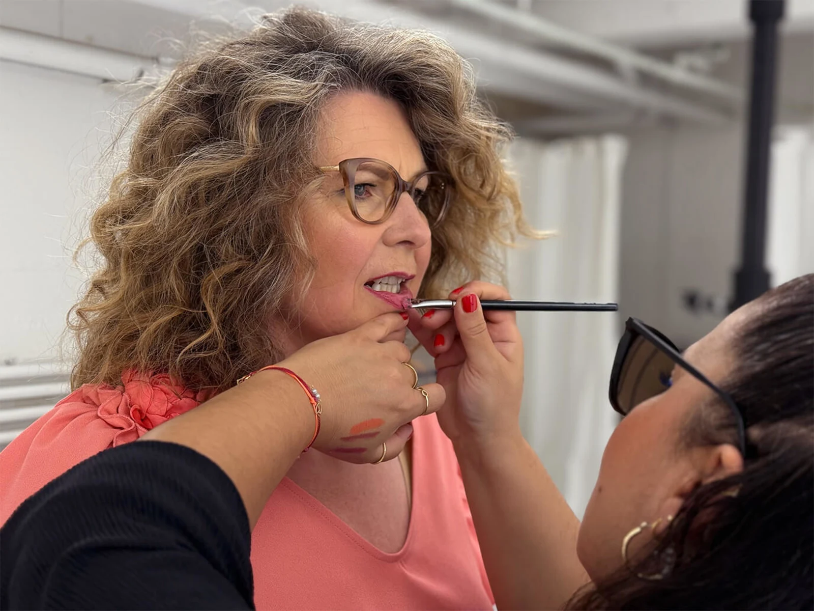 Eine Frau mit lockigem Haar wird geschminkt. Eine Person trägt Make Up auf das Gesicht der Frau auf. Der Hintergrund ist unscharf. Eine Frau mit lockigem Haar wird geschminkt. Eine Person trägt Make Up auf das Gesicht der Frau auf. Der Hintergrund ist unscharf.