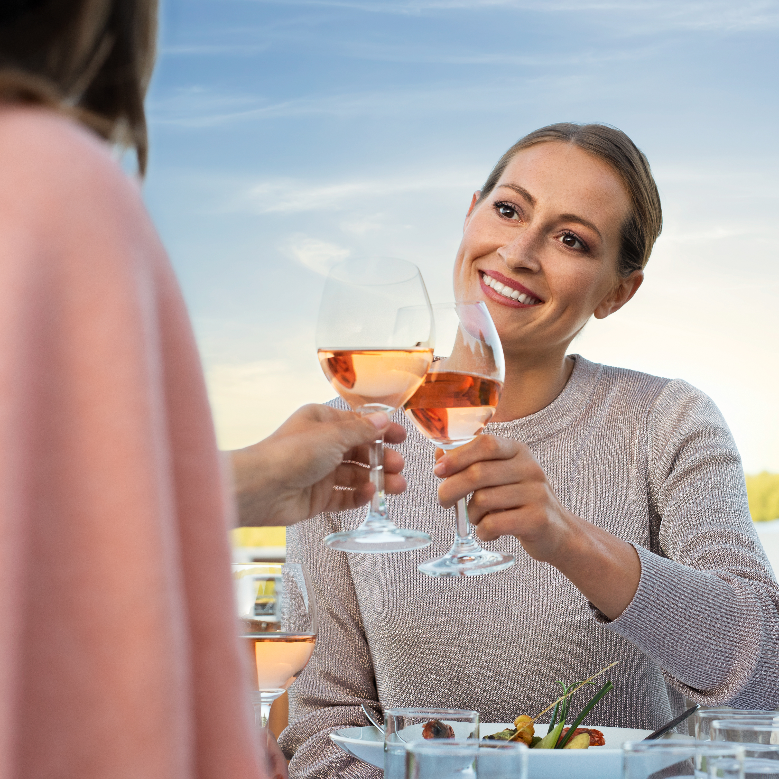 Zwei Models stoßen draußen mit Weingläsern an, ein gedeckter Tisch mit Speisen im Vordergrund.

