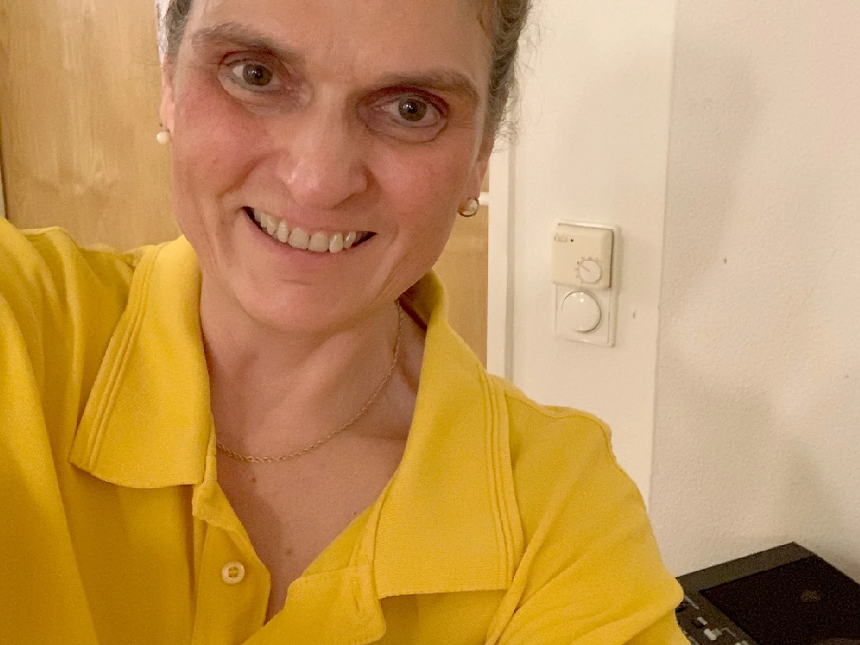 Eine Frau trägt ein gelbes Polohemd und hat eine Kette um den Hals. Im Hintergrund sind eine Wand und ein Lichtschalter sichtbar. Eine Frau trägt ein gelbes Polohemd und hat eine Kette um den Hals. Im Hintergrund sind eine Wand und ein Lichtschalter sichtbar.