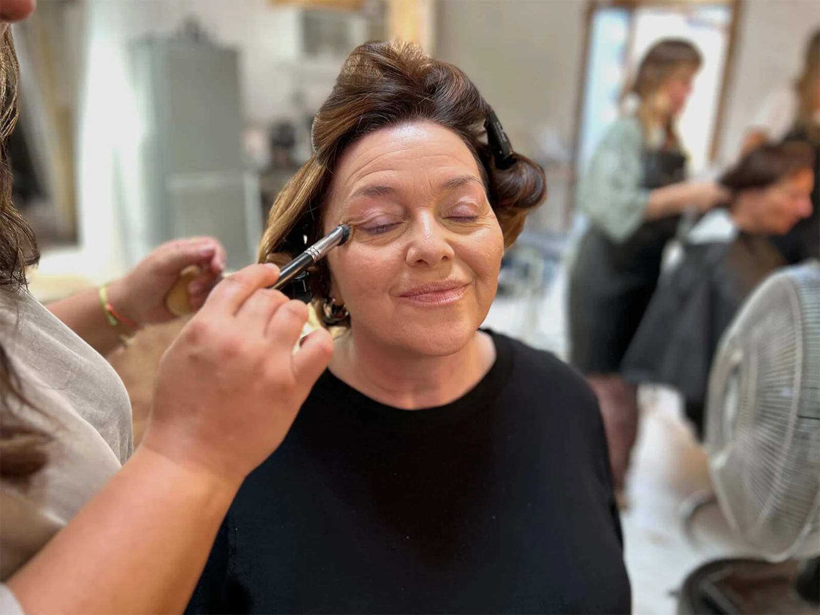Eine Frau erhält Make Up mit Pinsel, Nahaufnahme der Hände und des Gesichts, im Hintergrund weitere Personen in einem Friseursalon. Eine Frau erhält Make Up mit Pinsel, Nahaufnahme der Hände und des Gesichts, im Hintergrund weitere Personen in einem Friseursalon.