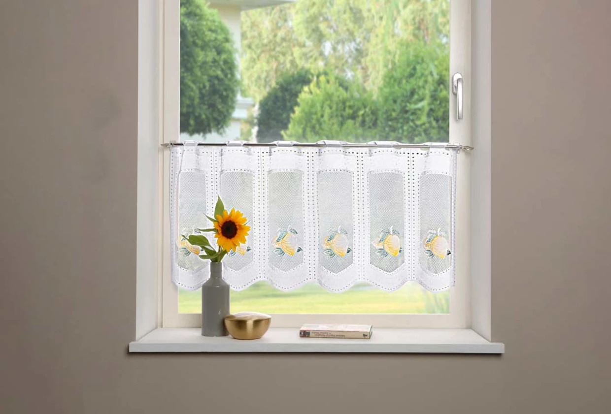 Fenster mit weißer Häkelgardine, verziert mit Zitronenmotiven, dekoriert mit einer Sonnenblume in einer Vase, einer goldenen Schale und einem Buch auf der Fensterbank. Fenster mit weißer Häkelgardine, verziert mit Zitronenmotiven, dekoriert mit einer Sonnenblume in einer Vase, einer goldenen Schale und einem Buch auf der Fensterbank.