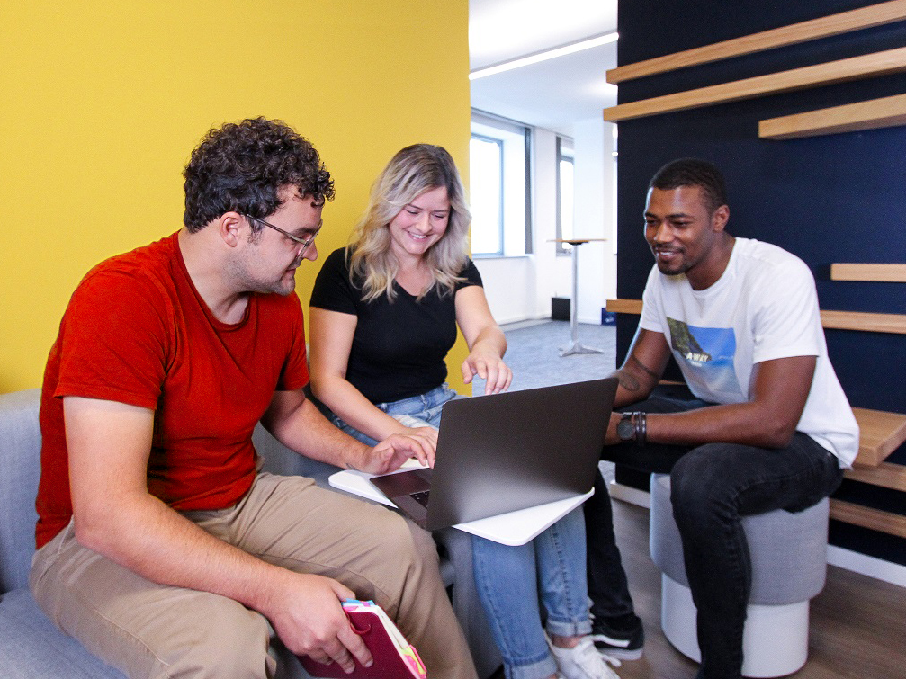 Drei Personen sitzen zusammen in einer modernen Büroumgebung vor einem Laptop und arbeiten gemeinsam, im Hintergrund eine gelbe Wand und Holzregale.