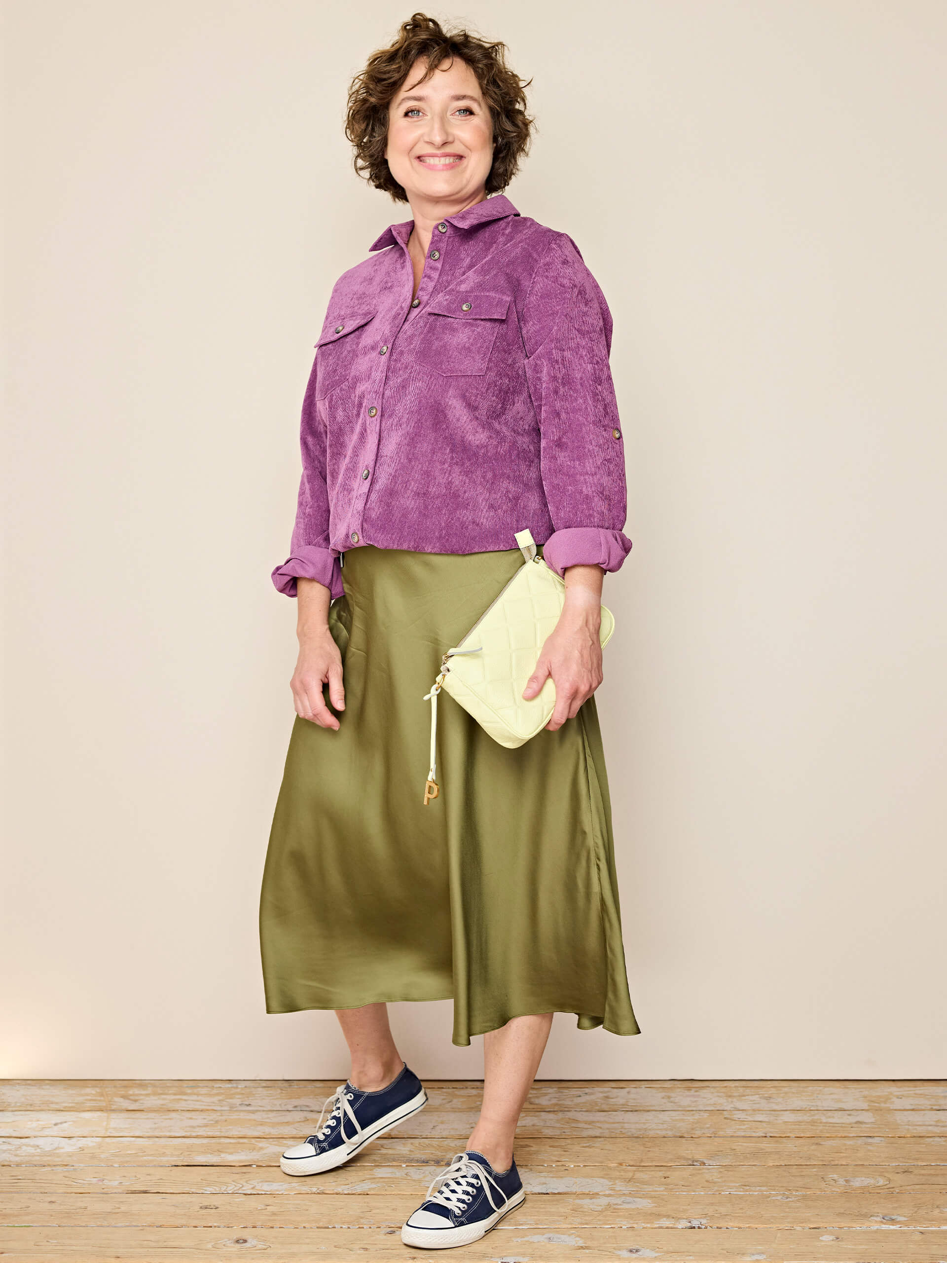 Ein Model trägt eine lila Cordjacke, einen olivgrünen Midirock, dunkelblaue Sneaker und hält eine helle Handtasche, vor beigem Hintergrund.