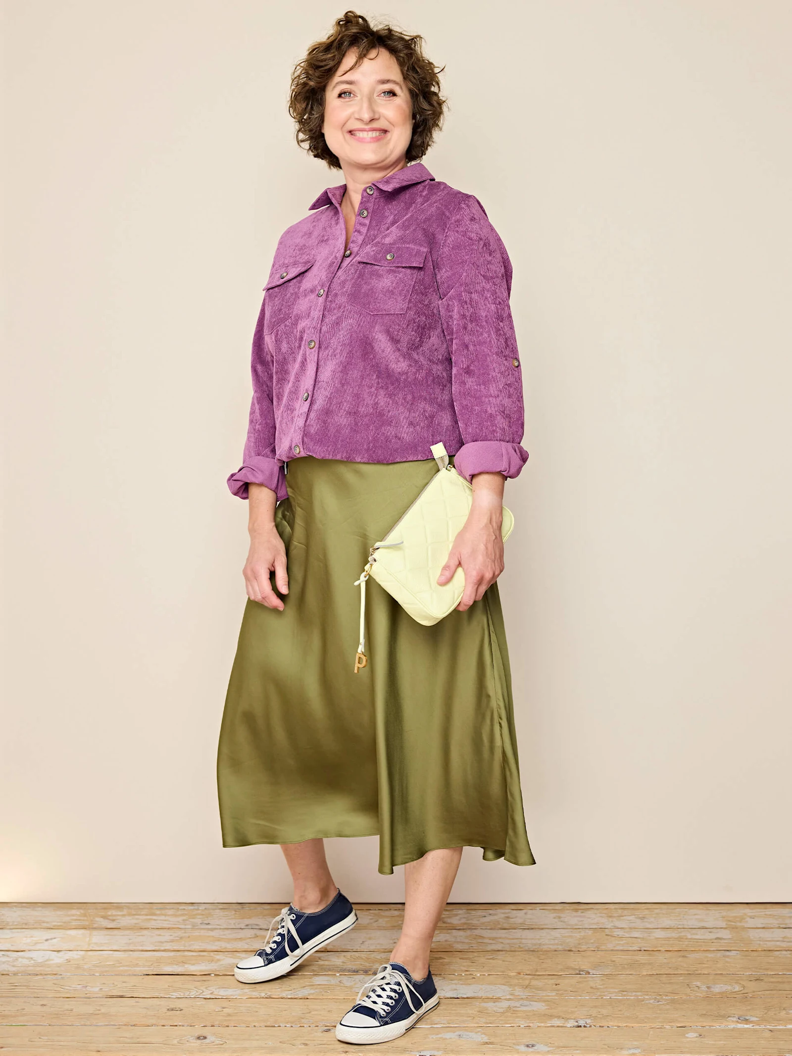 Ein Model trägt eine lila Cordjacke, einen olivgrünen Midirock, dunkelblaue Sneaker und hält eine helle Handtasche, vor beigem Hintergrund.