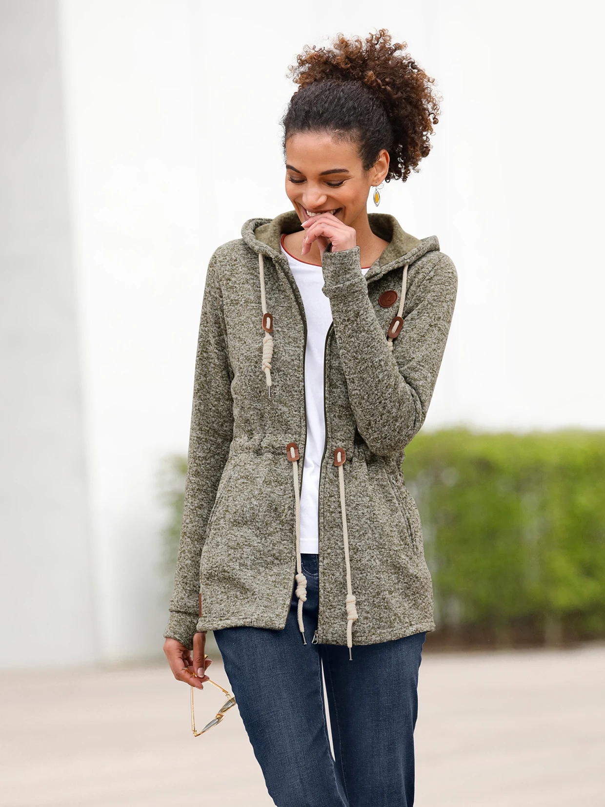 Damen-Outfit mit olivgrüner Strickjacke mit Kapuze und Kordelzug, weißem Shirt und blauer Jeans.