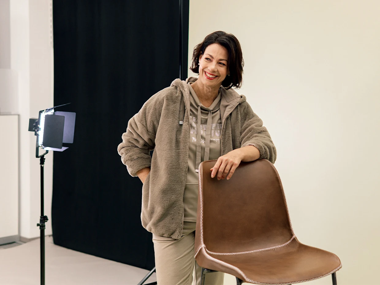 Eine Frau mit dunklen kurzen Haaren trägt eine schlammfarbene Fellimitatjacke, ein gleichfarbiges Oberteil und beige Hosen. Sie lehnt an einem Stuhl und posiert vor einer hellen Wand. Eine Frau mit dunklen kurzen Haaren trägt eine schlammfarbene Fellimitatjacke, ein gleichfarbiges Oberteil und beige Hosen. Sie lehnt an einem Stuhl und posiert vor einer hellen Wand.