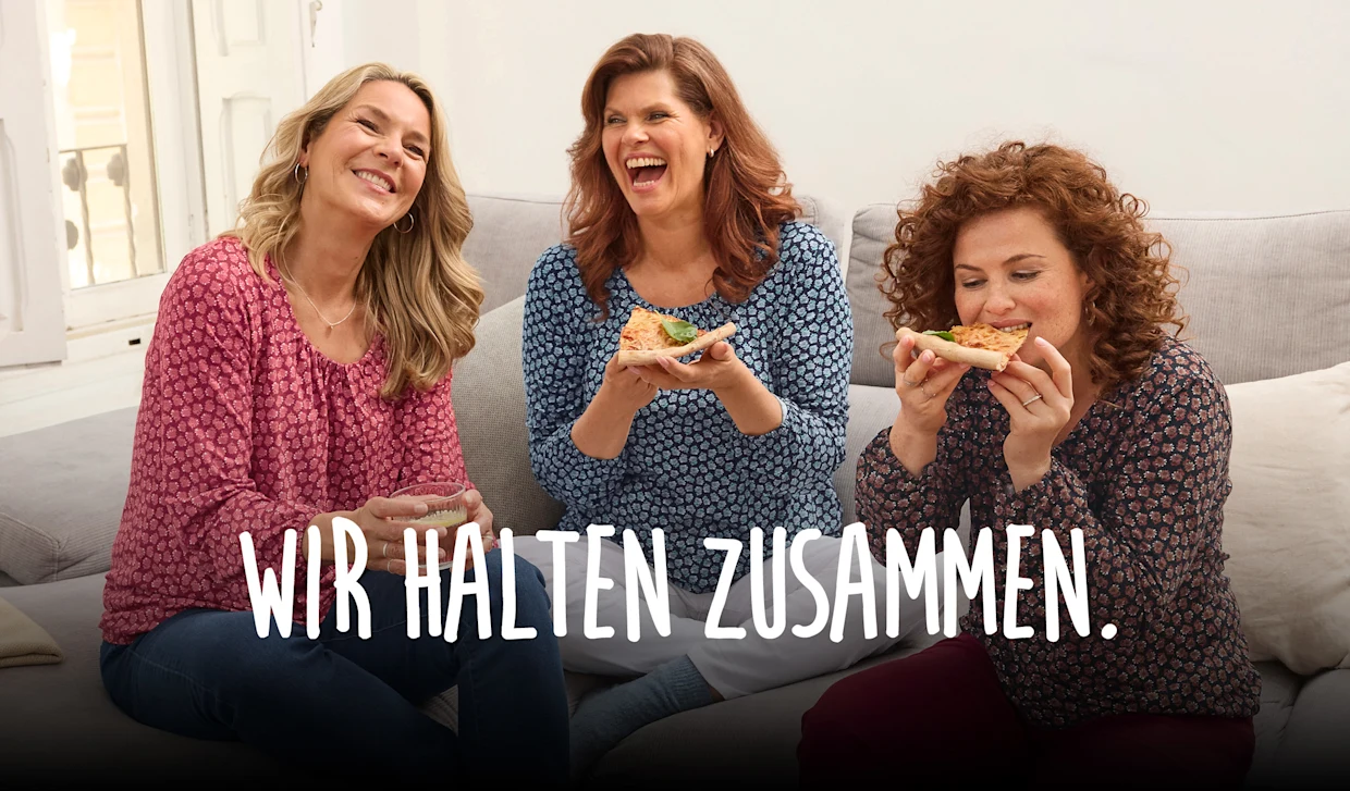 Eine Gruppe von Frauen sitzt lachend auf einem Sofa während sie Pizza essen. Auf dem Bild steht "Wir halten zusammen" geschrieben. Eine Gruppe von Frauen sitzt lachend auf einem Sofa während sie Pizza essen. Auf dem Bild steht "Wir halten zusammen" geschrieben.