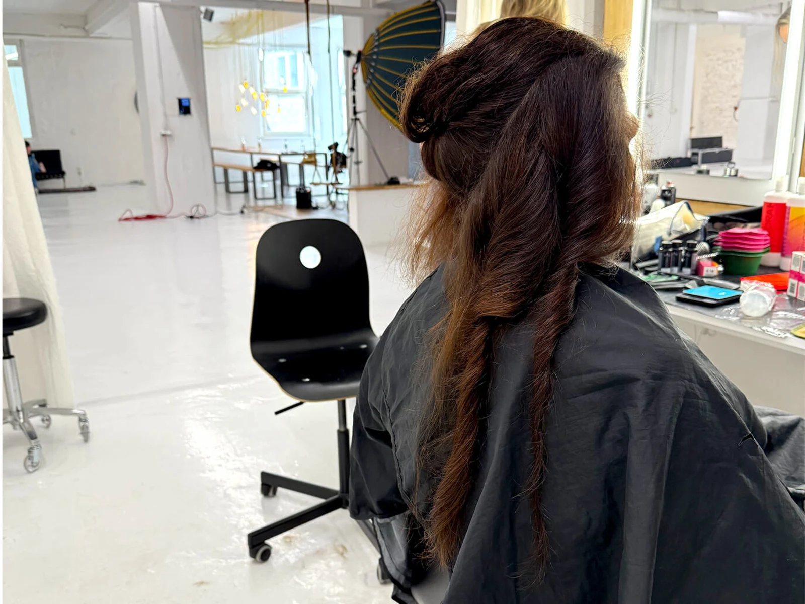 Eine Frau sitzt mit einem schwarzen Umhang im Friseurstuhl, während die Haare frisiert werden. Im Hintergrund sind verschiedene Utensilien sichtbar. Eine Frau sitzt mit einem schwarzen Umhang im Friseurstuhl, während die Haare frisiert werden. Im Hintergrund sind verschiedene Utensilien sichtbar.