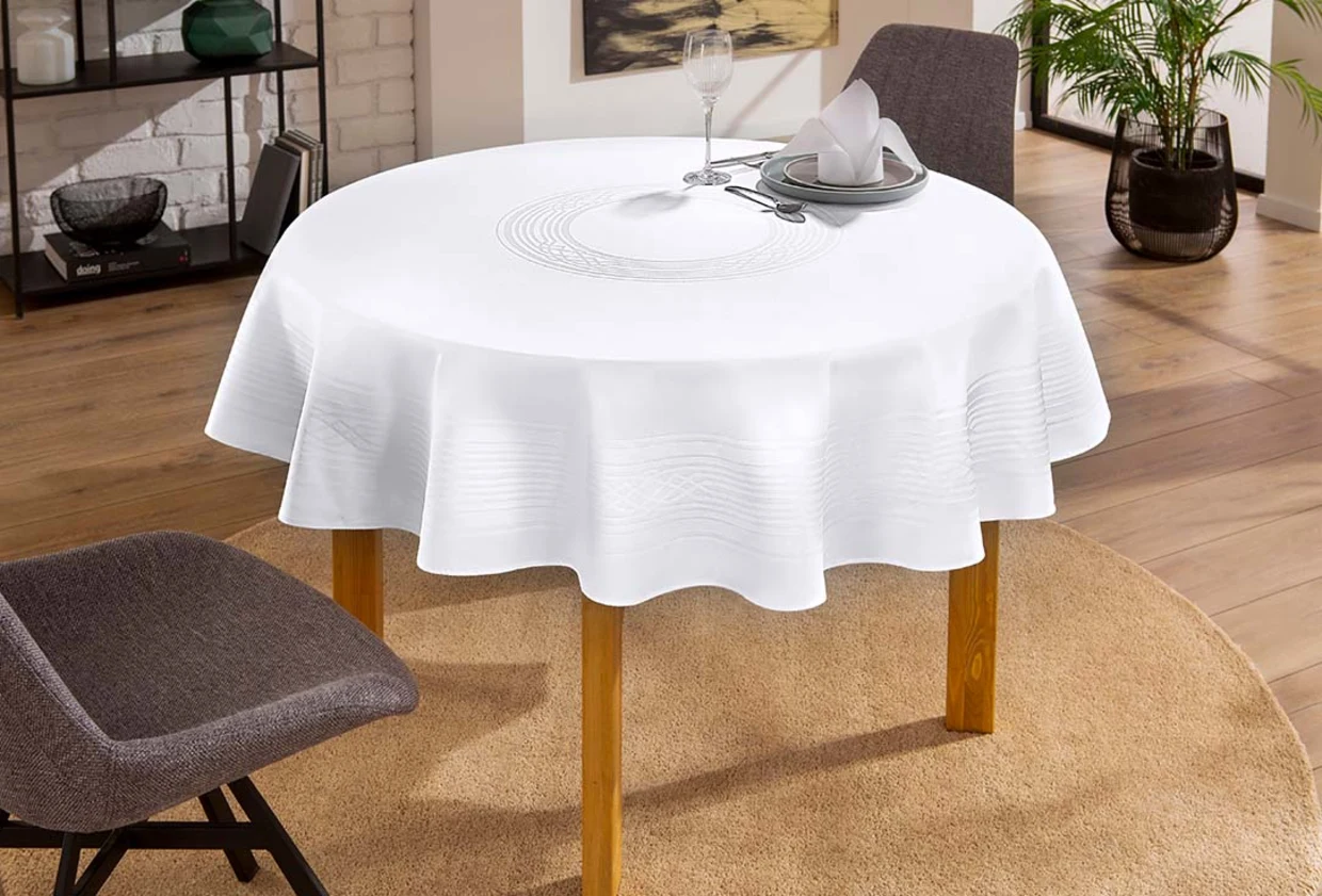 Runder Holztisch mit weißer Tischdecke in einem modern eingerichteten Raum, dekoriert mit einem gedeckten Teller, Besteck und Glas. Runder Holztisch mit weißer Tischdecke in einem modern eingerichteten Raum, dekoriert mit einem gedeckten Teller, Besteck und Glas.