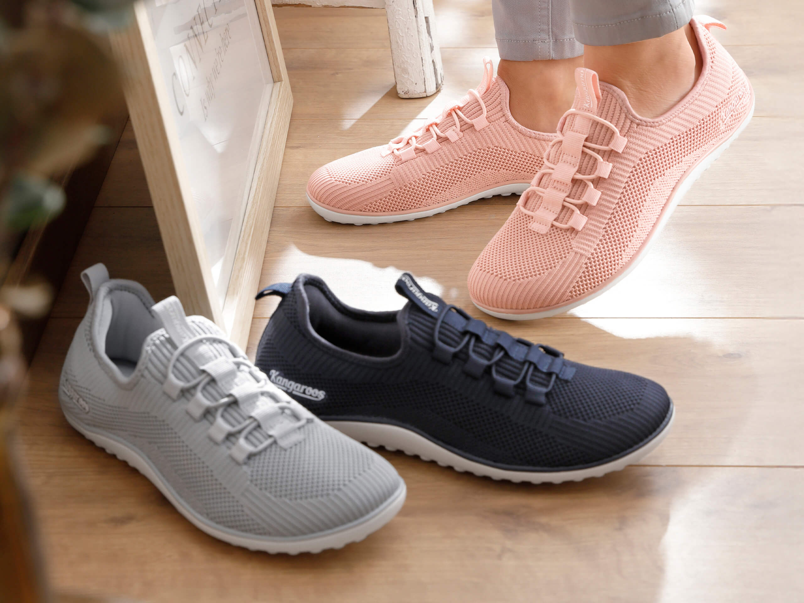 Eine Frau trägt hellrosé Sportschuhe aus Netzmaterial, davor stehen noch zwei Paar derselben Schuhe in den Farben grau und marine.