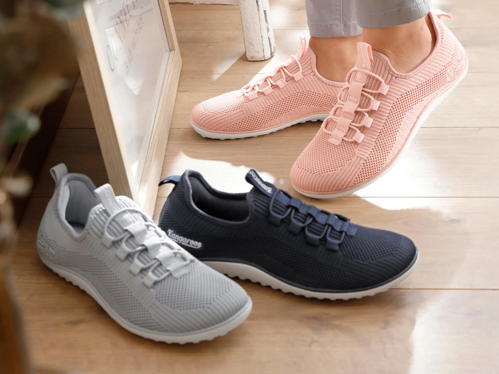 Eine Frau trägt hellrosé Sportschuhe aus Netzmaterial, davor stehen noch zwei Paar derselben Schuhe in den Farben grau und marine.