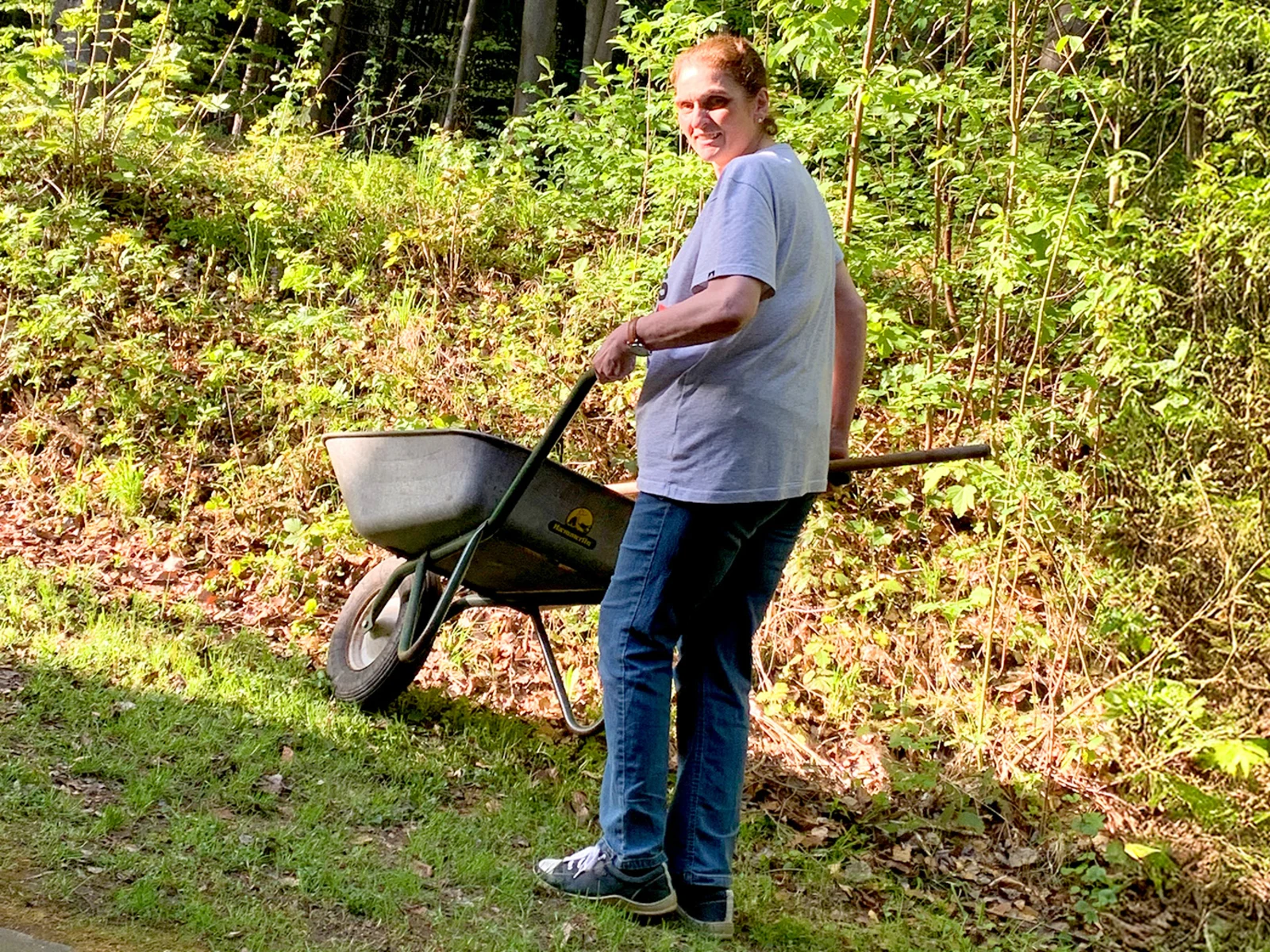 Eine Frau steht im Freien und schiebt eine Schubkarre. Sie trägt ein graues T-Shirt und eine blaue Jeans. Im Hintergrund sind grüne Pflanzen und Bäume sichtbar. Eine Frau steht im Freien und schiebt eine Schubkarre. Sie trägt ein graues T-Shirt und eine blaue Jeans. Im Hintergrund sind grüne Pflanzen und Bäume sichtbar.