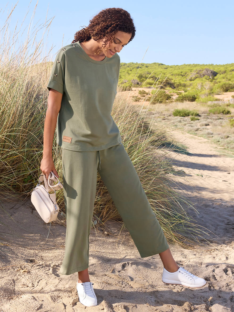 Ein Model trägt ein olivgrünes, lockeres T-Shirt, eine olivgrüne weite Hose und weiße Sneakers, steht auf sandigem Boden mit Gras und hält eine kleine helle Tasche.