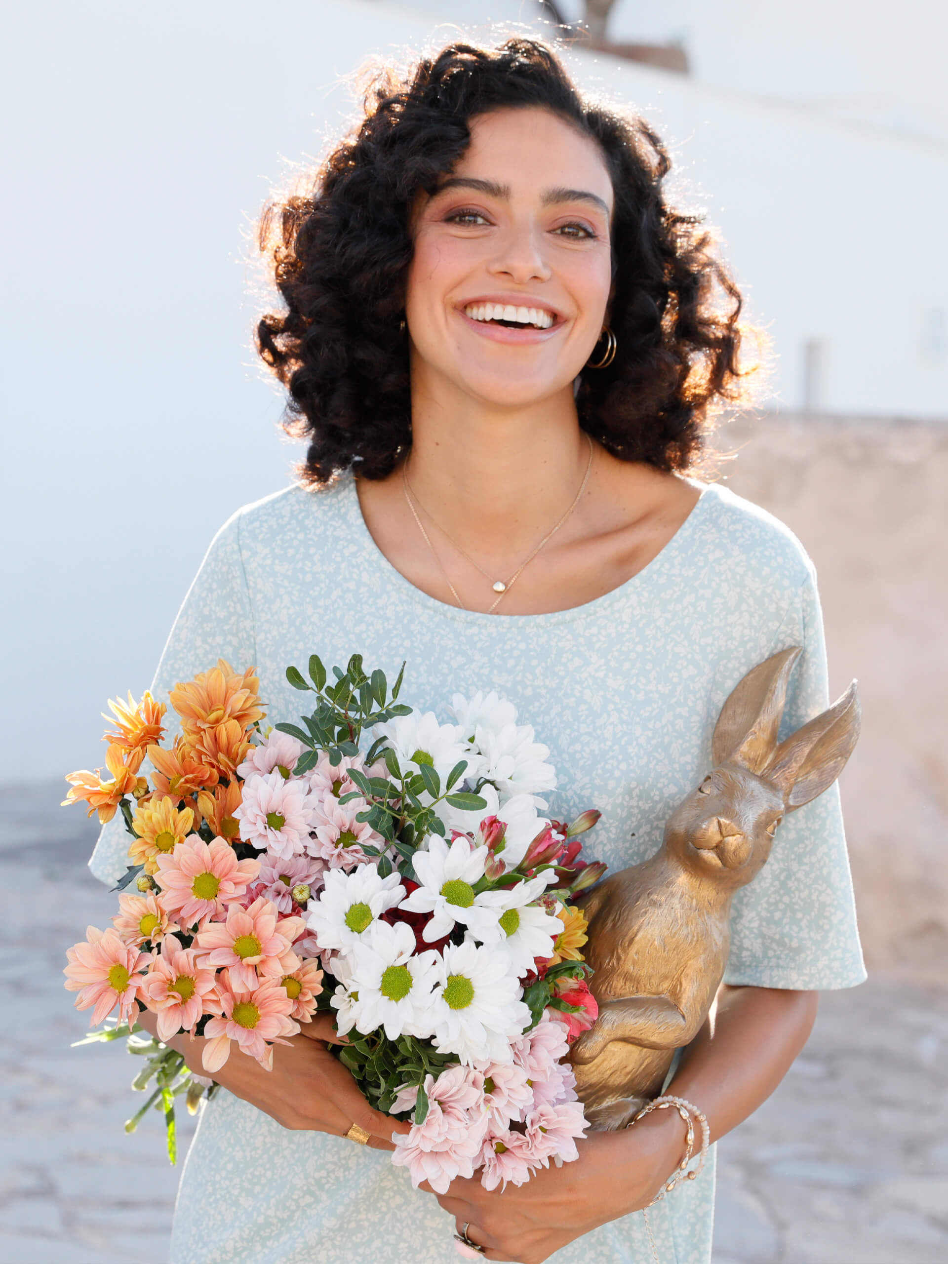Ein Model trägt ein hellblau-weiß gemustertes Kleid mit kurzen Ärmeln, hält einen bunten Blumenstrauß und eine goldene Hasenfigur im Arm.