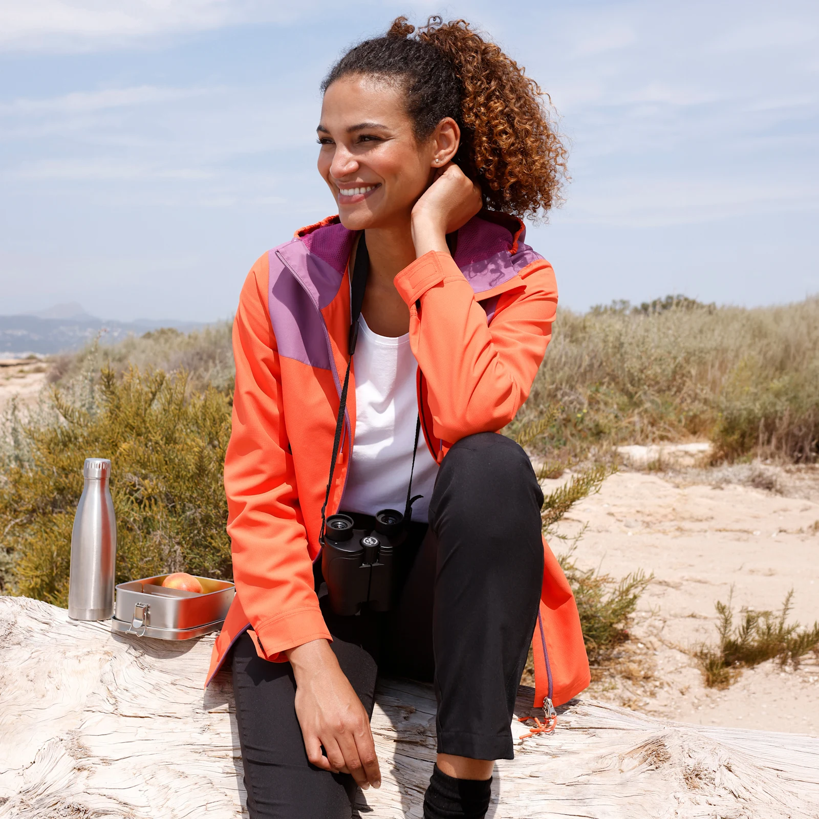 Ein Model sitzt draußen auf einem Felsen, trägt eine orangefarbene Outdoorjacke, schwarze Hose und Sportschuhe, Fernglas um den Hals, Trinkflasche und Dose daneben.