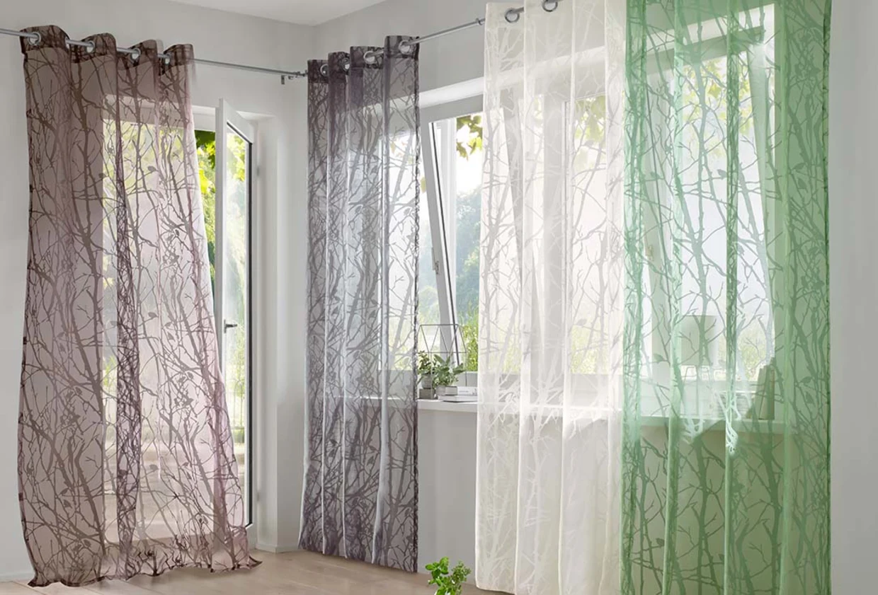 Transparente Vorhänge mit Ast-Muster in Braun, Grau, Weiß und Grün vor großen Fenstern in einem hellen Raum. Transparente Vorhänge mit Ast-Muster in Braun, Grau, Weiß und Grün vor großen Fenstern in einem hellen Raum.