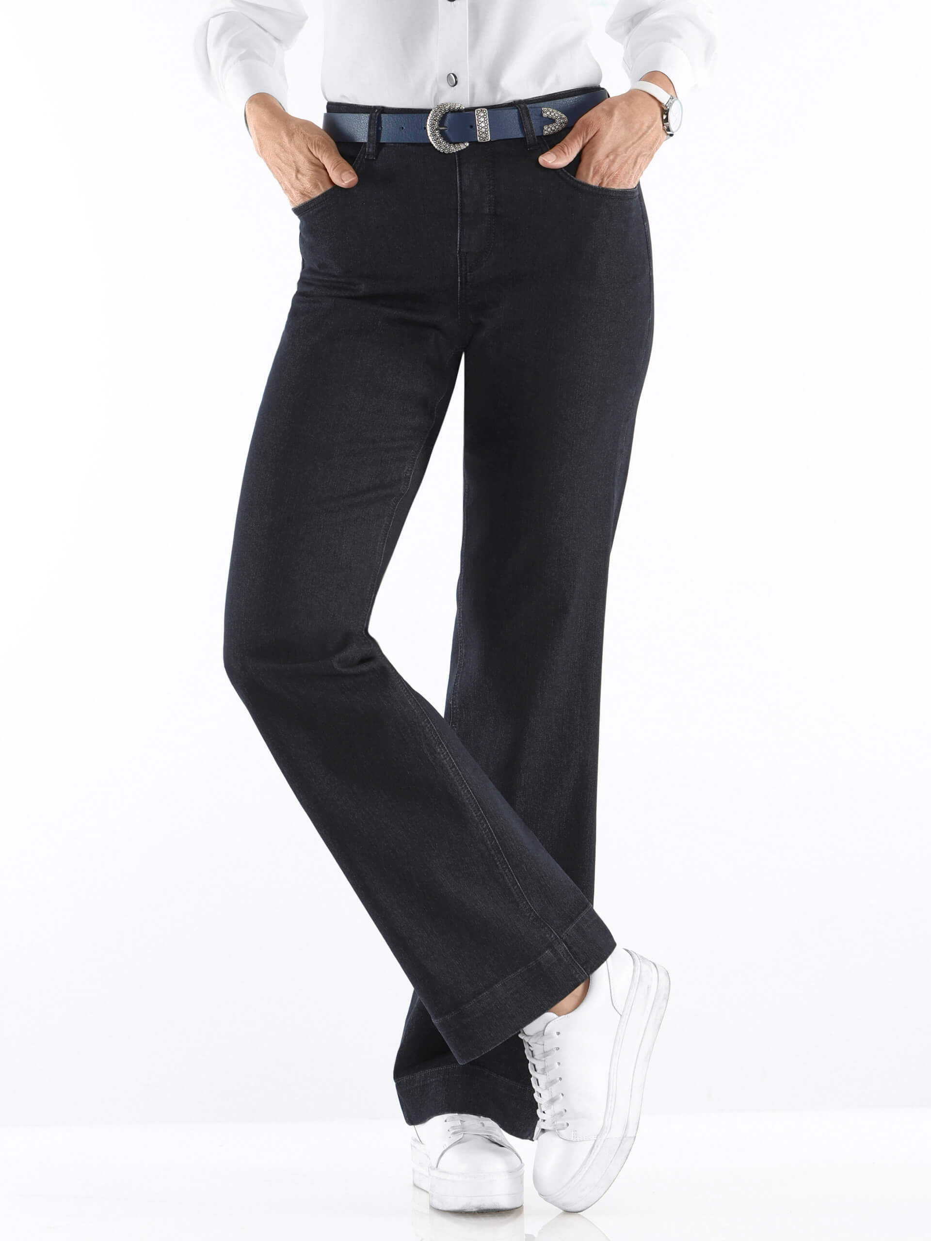 Ein Model trägt schwarze Jeans und ein weißes Oberteil, kombiniert mit einem schwarzen Gürtel und weißen Sneakers.