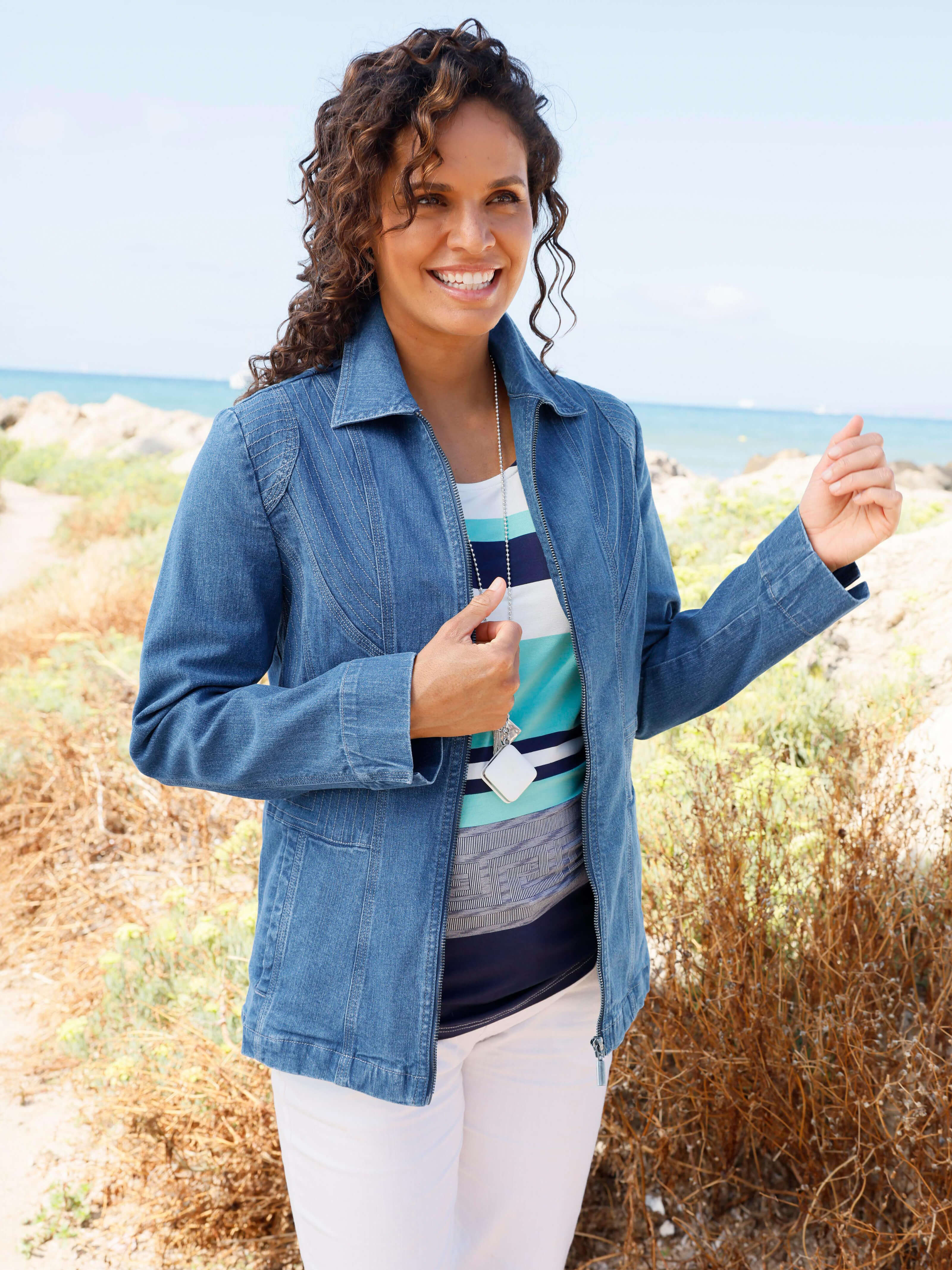 Ein Model trägt eine blaue Jeansjacke über einem gemusterten Oberteil und steht an einem Küstenweg mit Blick aufs Meer.