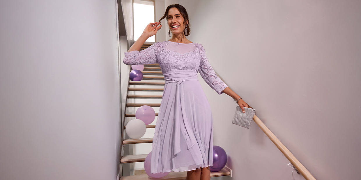 Ein weibliches Model trägt ein fliederfarbenes, knielanges Kleid mit 3/4-Ärmeln, Spitze und Glitzersteinchen, hält eine silberne Clutch auf einer Treppe mit Luftballons.