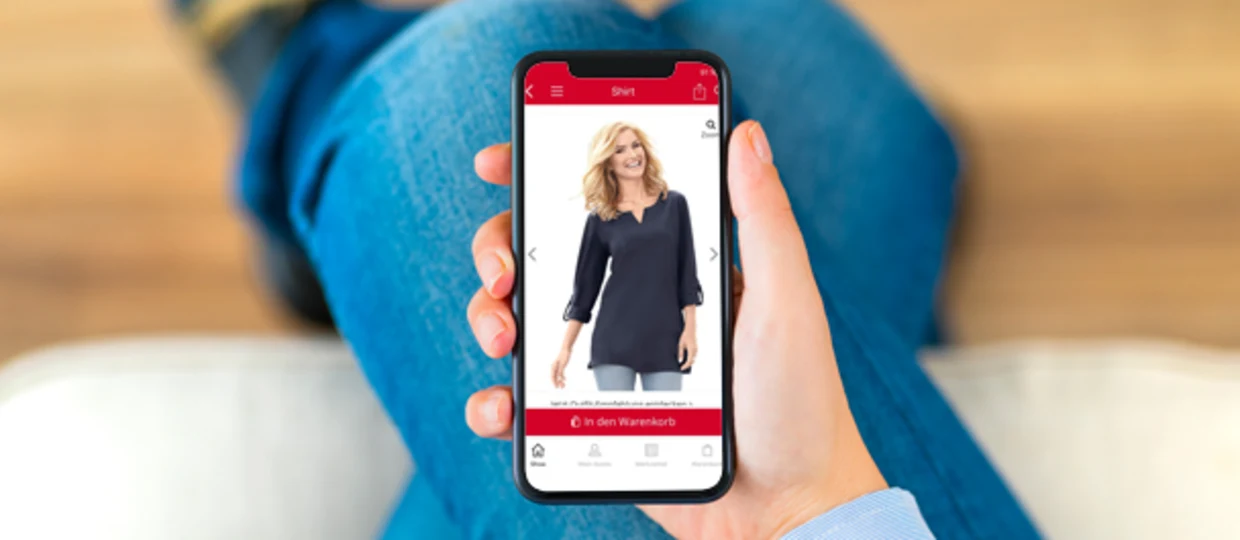 Nahaufnahme einer Hand, die ein Smartphone hält, auf dem ein Online-Shop geöffnet ist. Der Bildschirm zeigt ein dunkelblaues Damen-Shirt mit Dreiviertelärmeln und die Option "In den Warenkorb". Nahaufnahme einer Hand, die ein Smartphone hält, auf dem ein Online-Shop geöffnet ist. Der Bildschirm zeigt ein dunkelblaues Damen-Shirt mit Dreiviertelärmeln und die Option "In den Warenkorb".