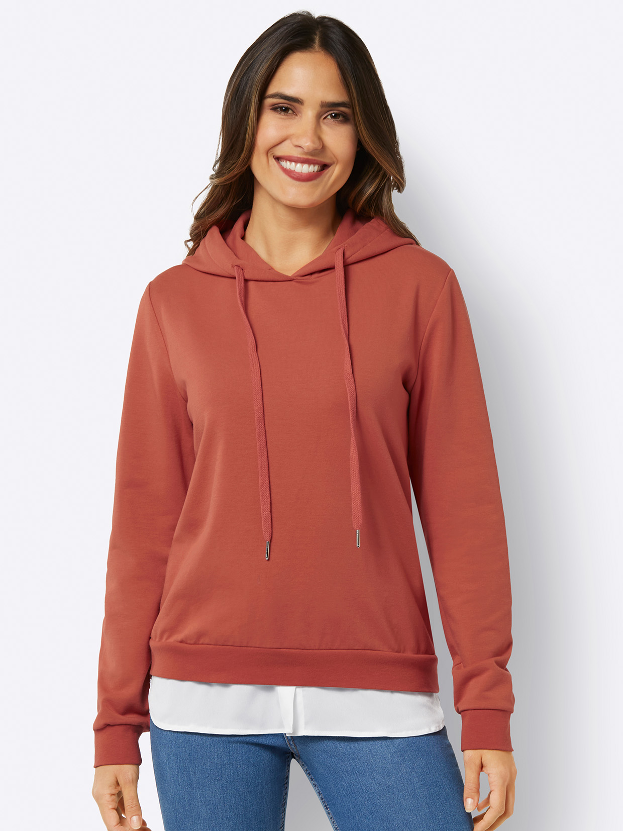 Damen-Hoodie in Terrakotta mit Kapuze, Kordelzug und weißem Schicht-Effekt, kombiniert mit einer blauen Jeans.