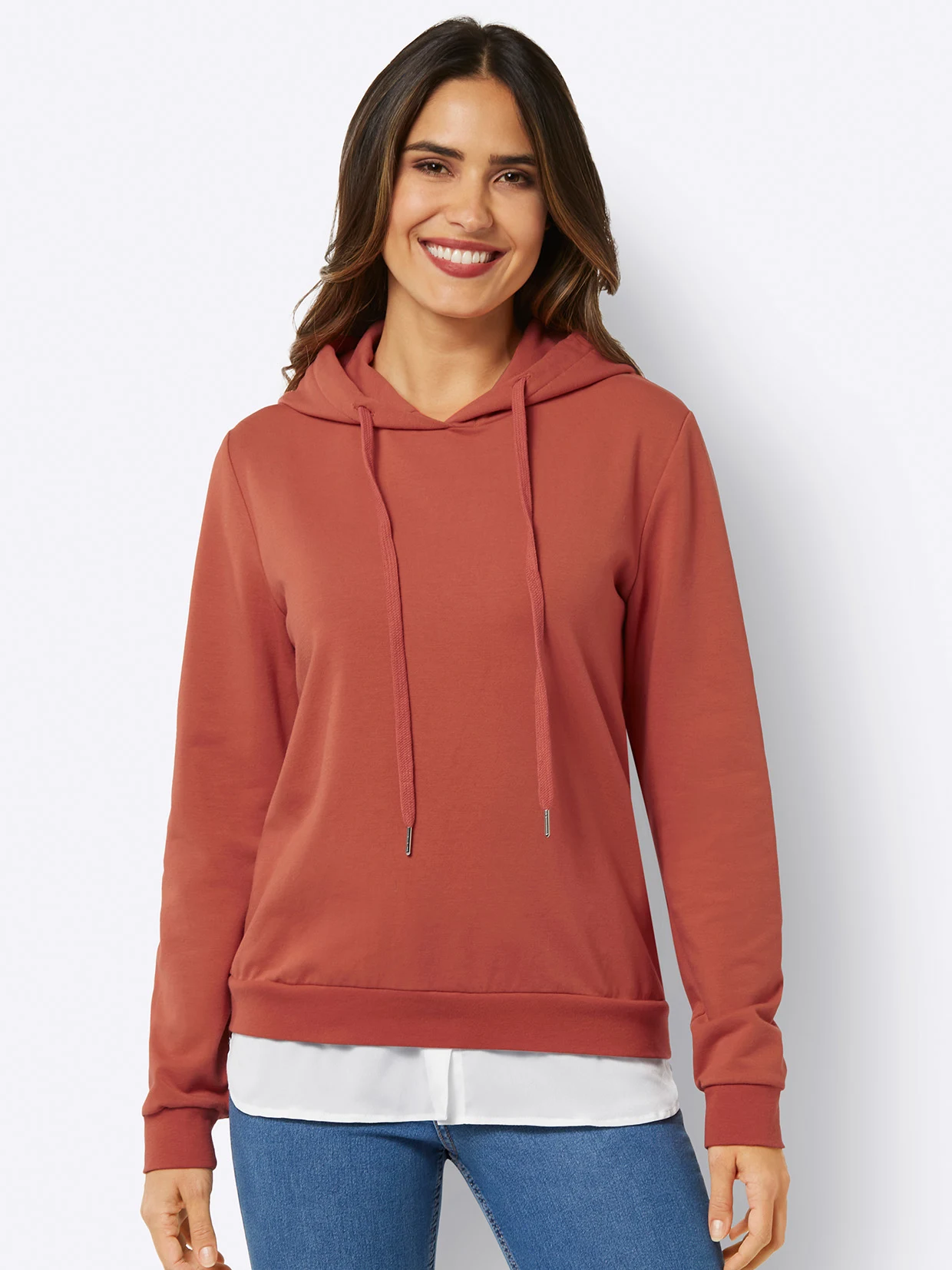 Damen-Hoodie in Terrakotta mit Kapuze, Kordelzug und weißem Schicht-Effekt, kombiniert mit einer blauen Jeans.