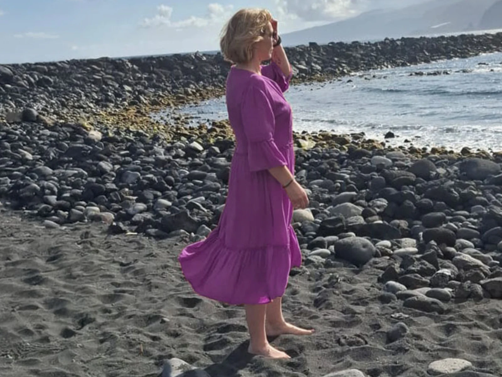 Eine Frau steht barfuß am Strand, trägt ein lila Kleid und hält sich die Haare aus dem Gesicht. Im Hintergrund sind Steine und das Meer sichtbar. Eine Frau steht barfuß am Strand, trägt ein lila Kleid und hält sich die Haare aus dem Gesicht. Im Hintergrund sind Steine und das Meer sichtbar.