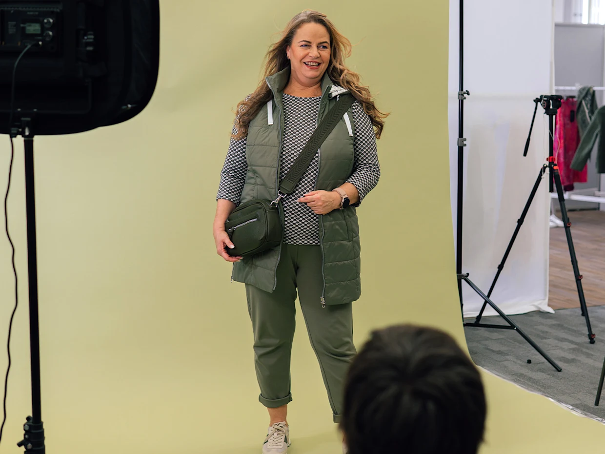 Eine blonde Frau trägt ein khakifarbenes Outfit und posiert vor einem gelben Studiohintergrund während ein Fotograf im Vordergrund mit dem Rücken zur Kamera sitzt, den Blick auf die Frau gerichtet. Eine blonde Frau trägt ein khakifarbenes Outfit und posiert vor einem gelben Studiohintergrund während ein Fotograf im Vordergrund mit dem Rücken zur Kamera sitzt, den Blick auf die Frau gerichtet.