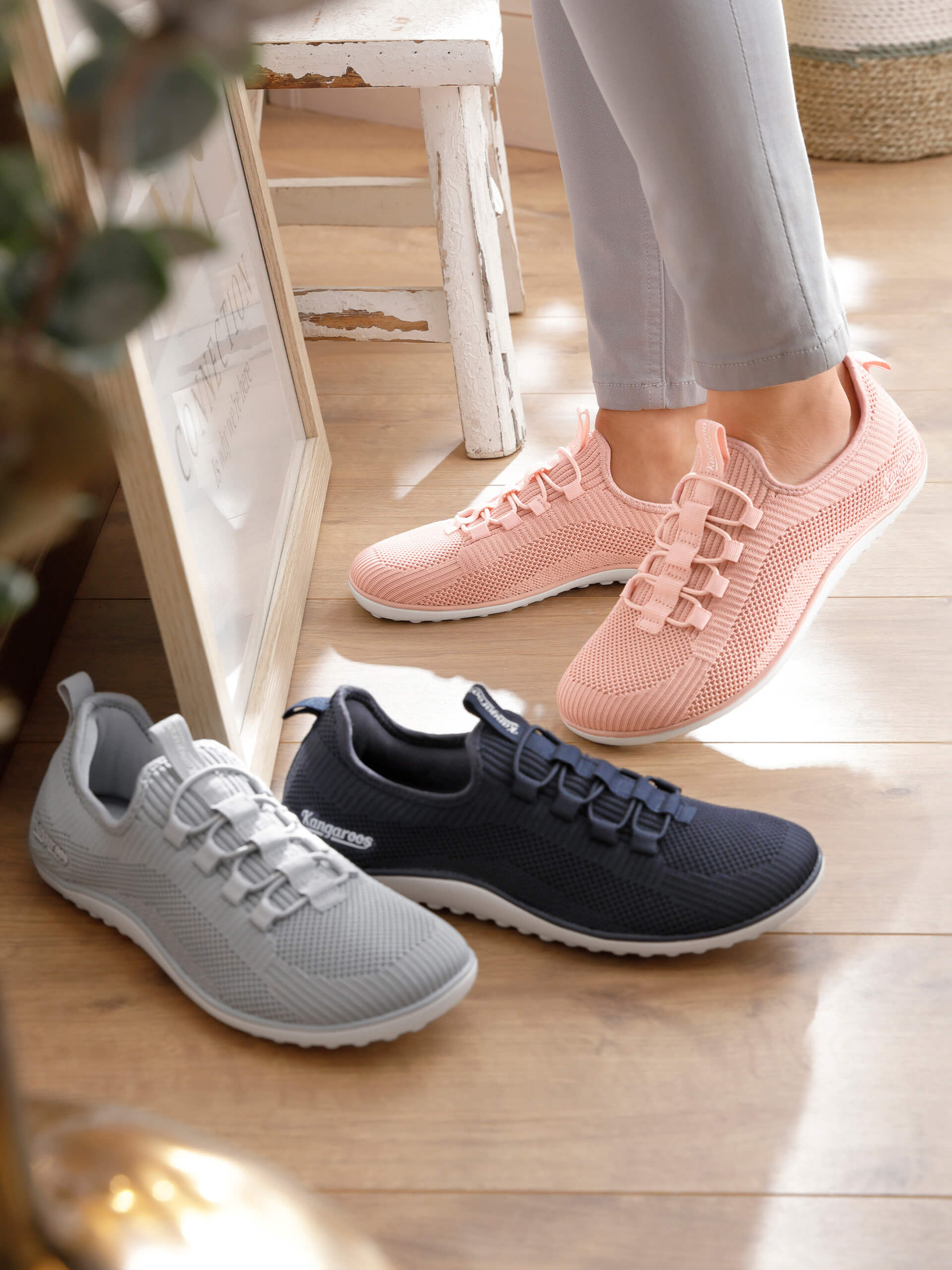 Eine Frau trägt hellrosé Sportschuhe aus Netzmaterial, davor stehen noch zwei Paar derselben Schuhe in den Farben grau und marine.