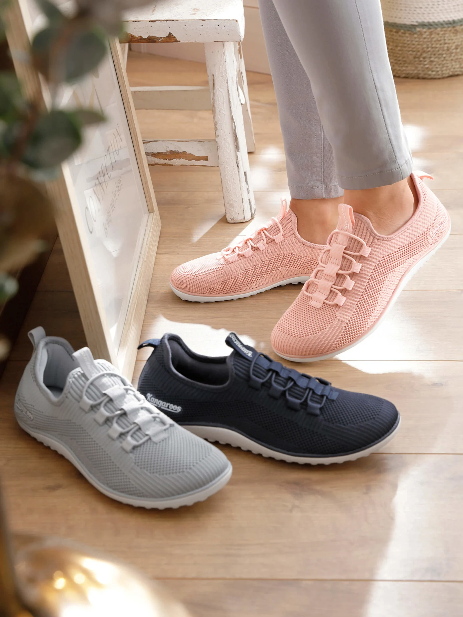 Eine Frau trägt hellrosé Sportschuhe aus Netzmaterial, davor stehen noch zwei Paar derselben Schuhe in den Farben grau und marine.