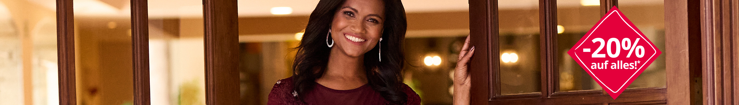 Das Bild zeigt eine Frau in einem bordeauxroten Kleid, die vor einer geöffneten Holztür steht. Im Hintergrund ist ein warm beleuchtetes Interieur zu sehen. Der Text befindet sich in einem roten Rautensymbol. Text: '-20% auf alles!*‘.