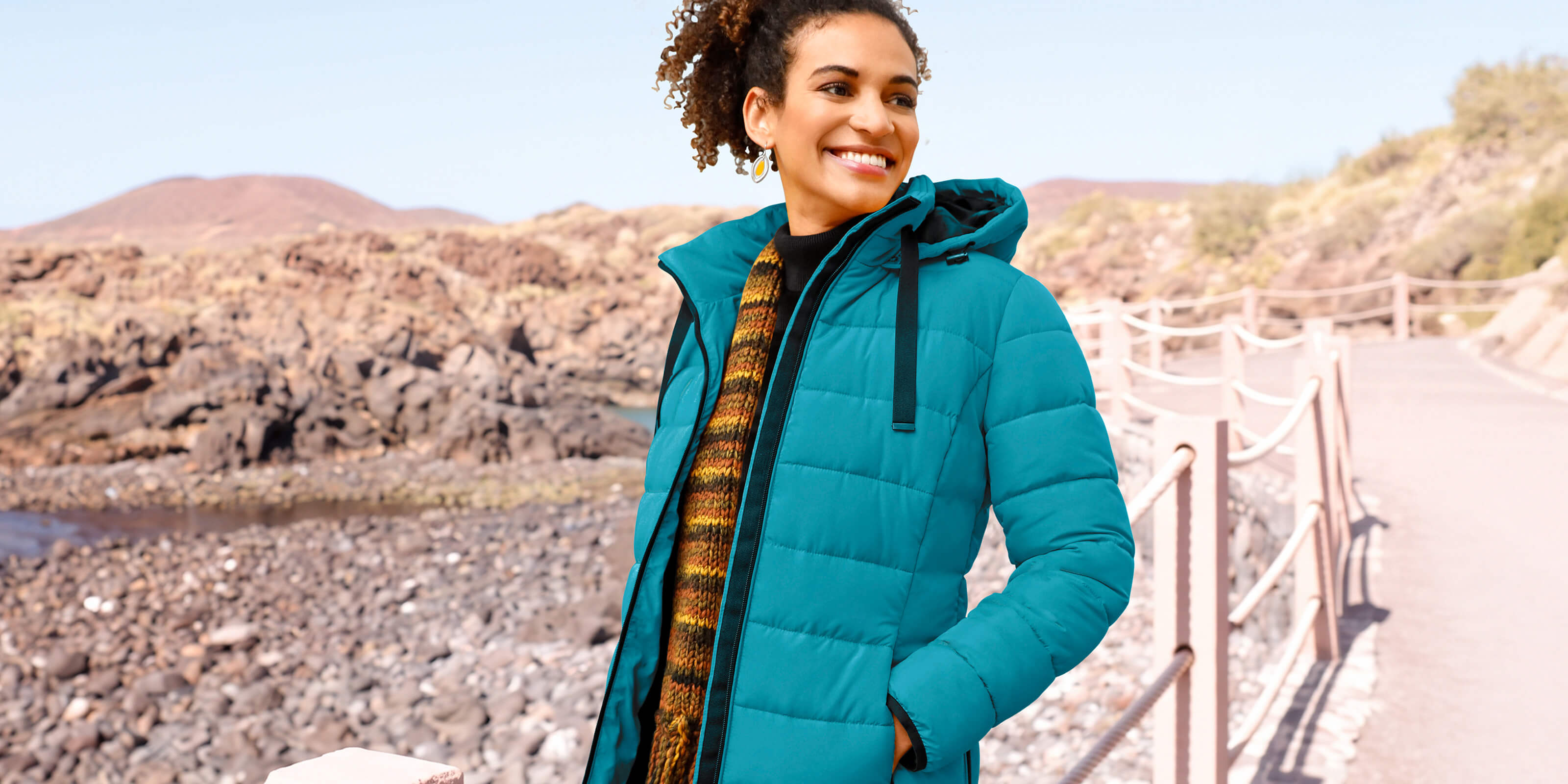 Eine lächelnde Frau steht im Freien und trägt eine offene, türkisfarbene Steppjacke mit Kapuze, einen schwarzen Rollkragenpullover und einen bunten Strickschal.