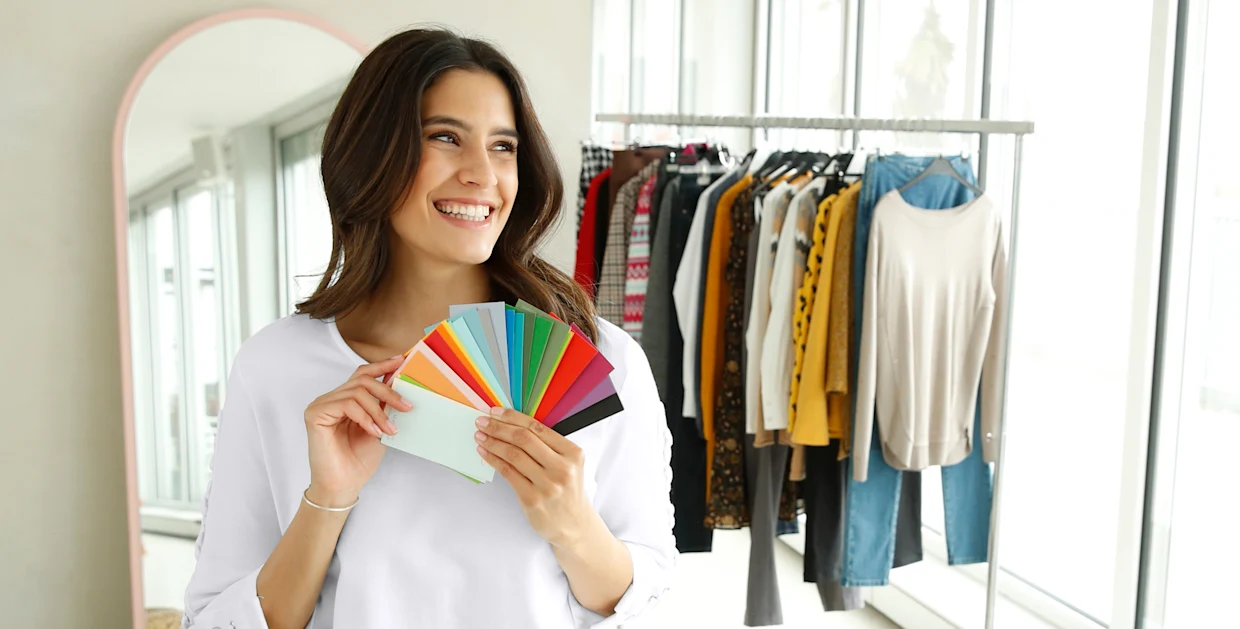 Vrouw houdt een waaier met kleurstalen vast voor een kledingrek met diverse outfits. Vrouw houdt een waaier met kleurstalen vast voor een kledingrek met diverse outfits.