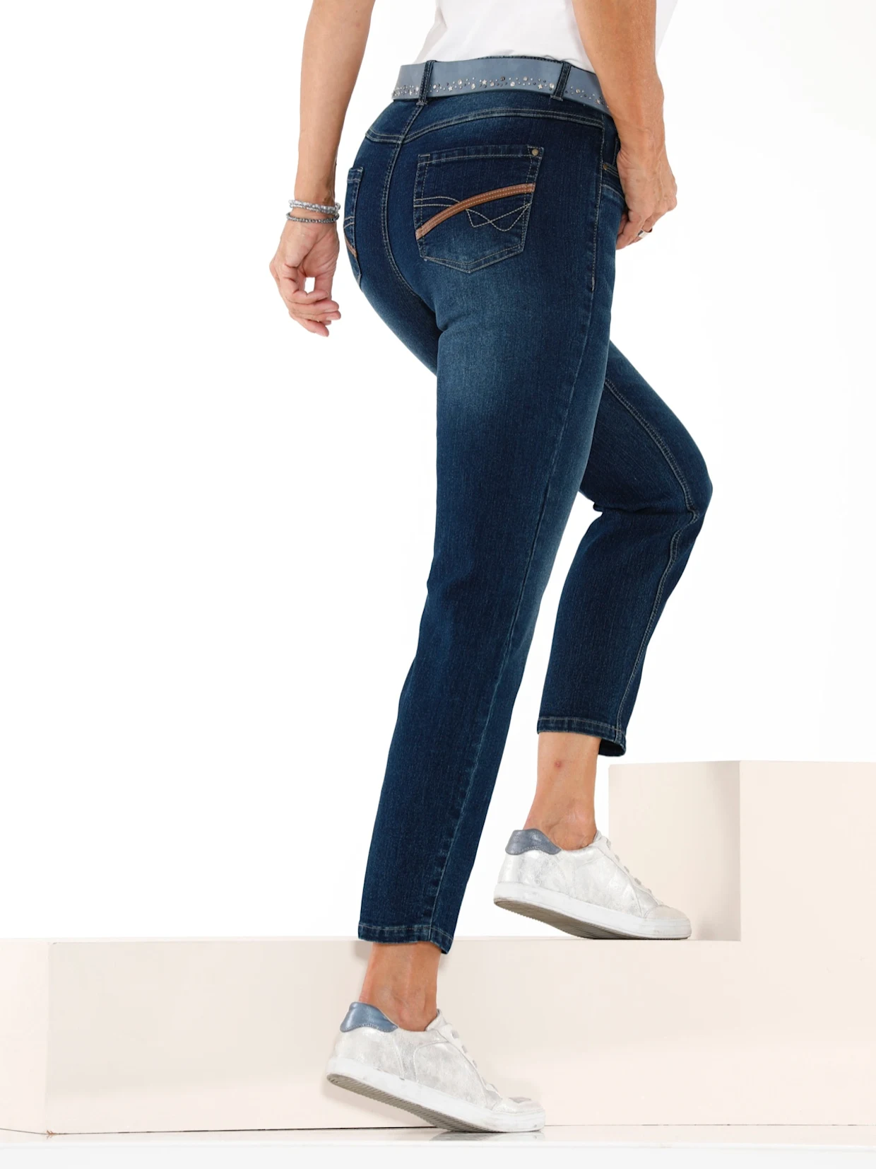 Frau in dunkelblauer Jeans mit dekorativen Gesäßtaschen und weiß-blauen Sneakers steigt eine helle Treppe hinauf.